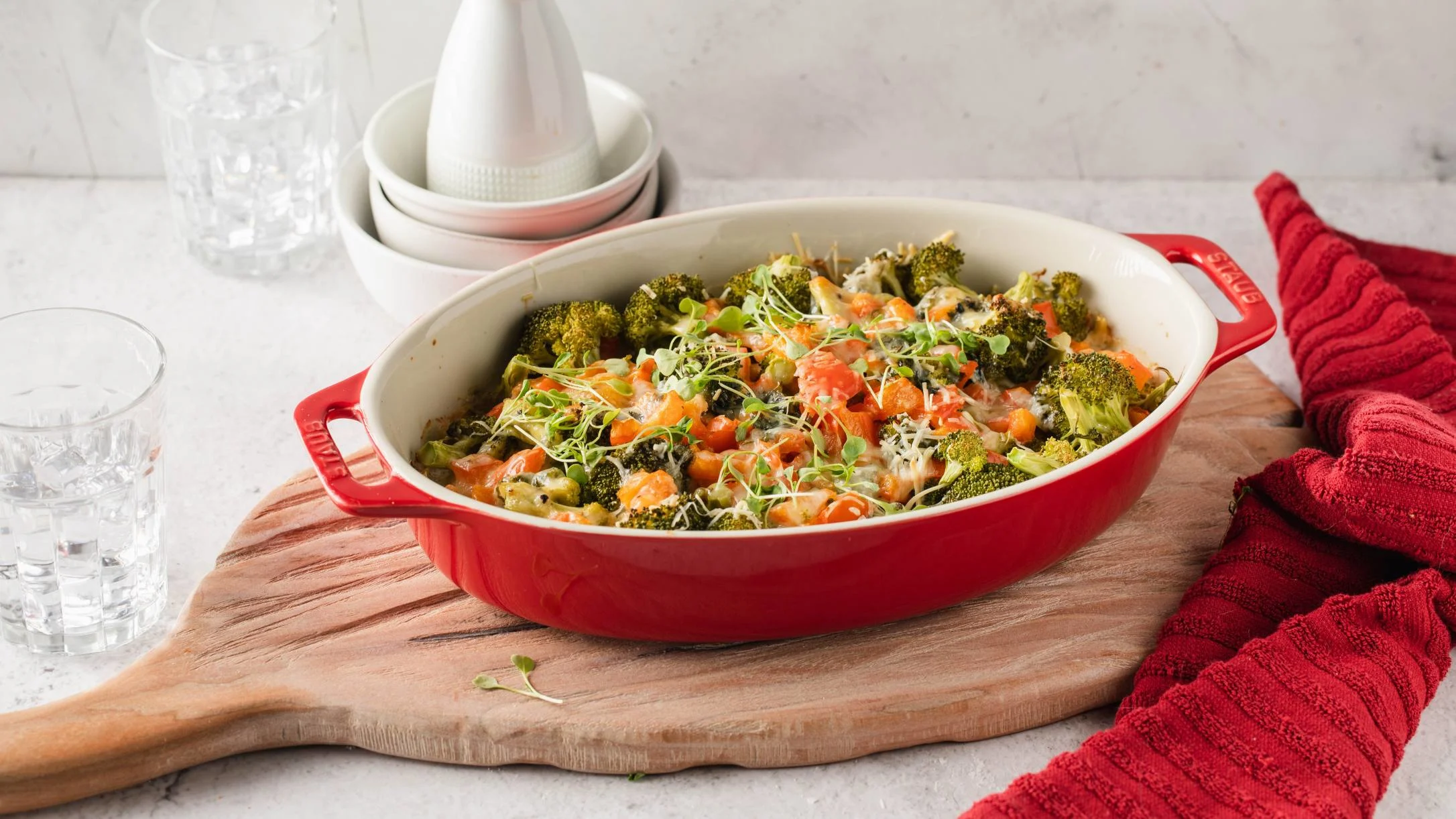 Baked chicken casserole with broccoli and carrots in a red dish, served on wooden board with water glasses nearby.