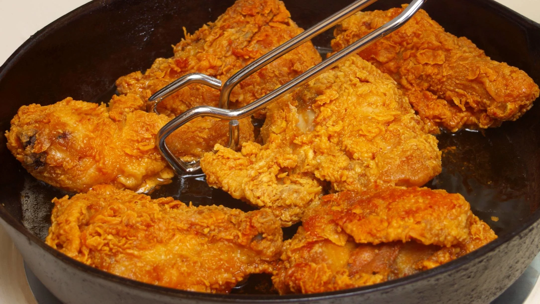 Golden-brown fried chicken pieces cooking in a black cast iron skillet with metal tongs resting on top.