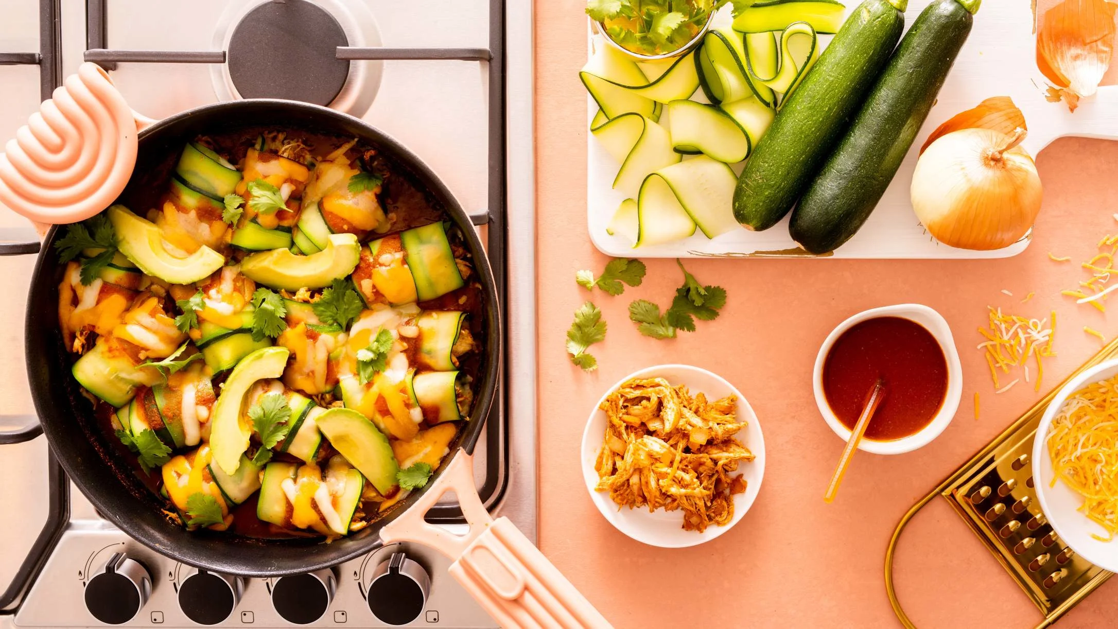 Cooking scene with avocado and zucchini dish in pan, fresh ingredients and sauce bowls on peach-colored countertop.