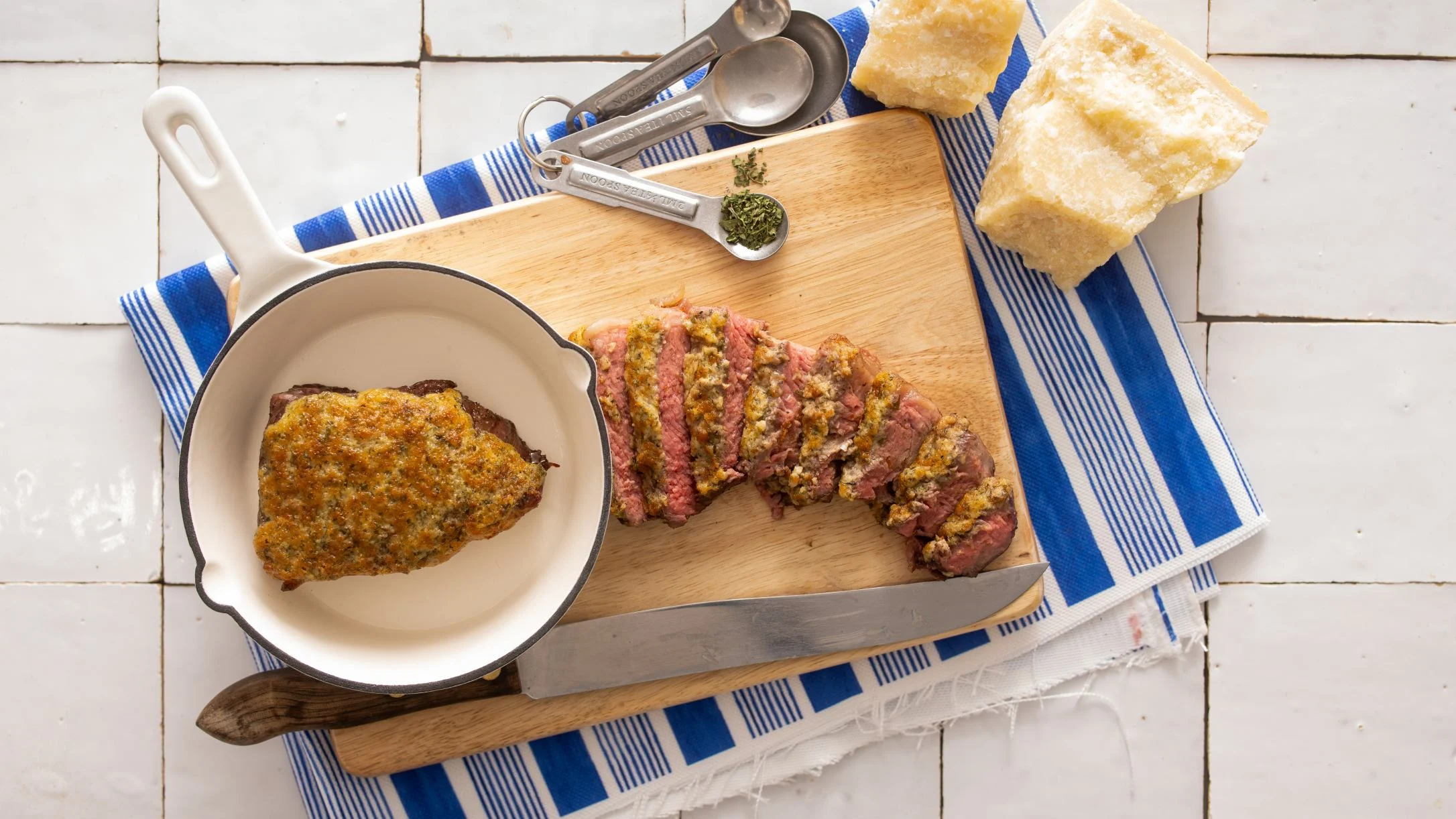 Sliced herb-crusted steak on a wooden cutting board with a knife, small skillet, measuring spoons, and Parmesan cheese on a blue striped towel.