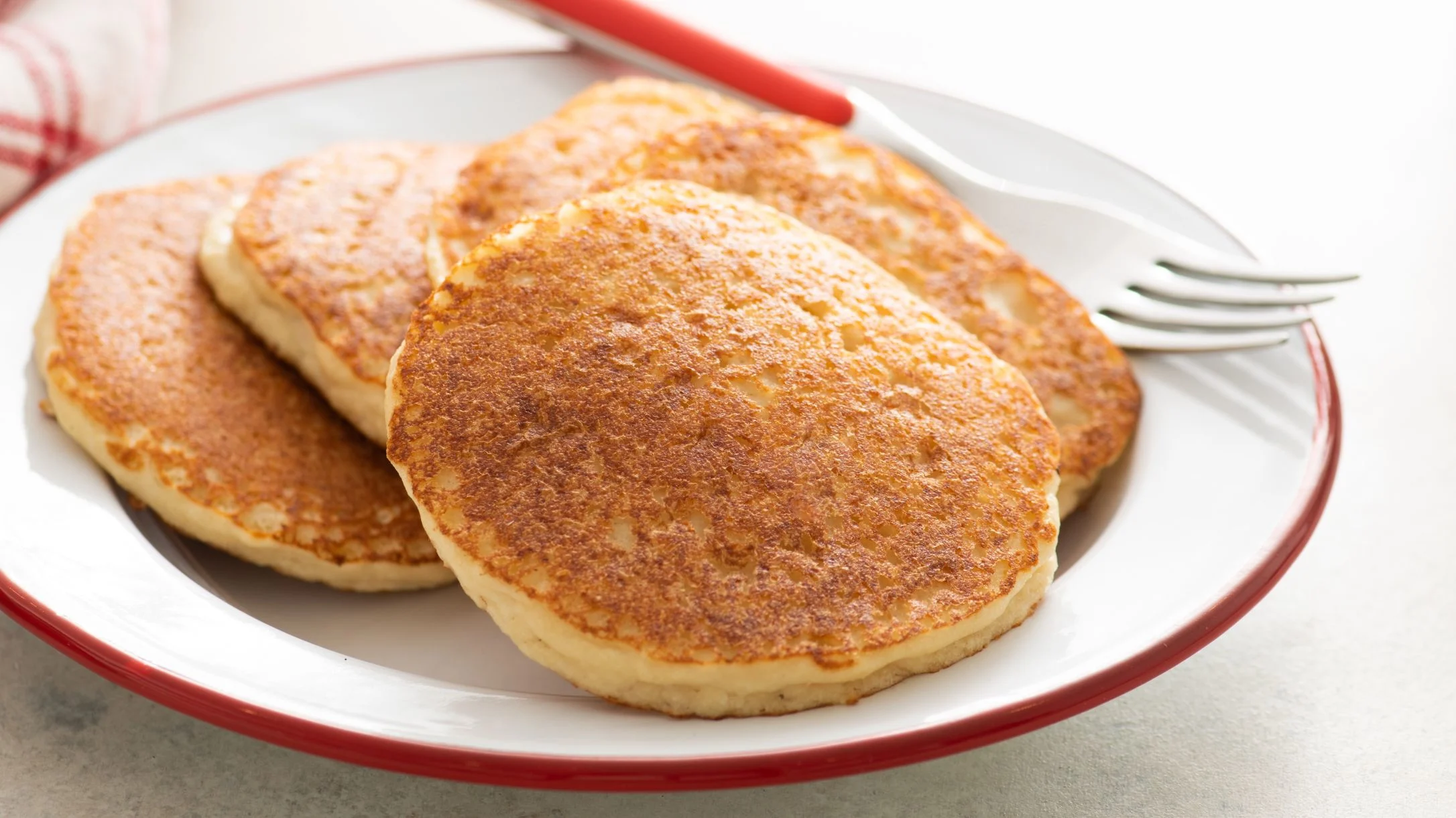 Four golden-brown pancakes stacked on a white plate with a red rim, accompanied by a fork with a red handle.