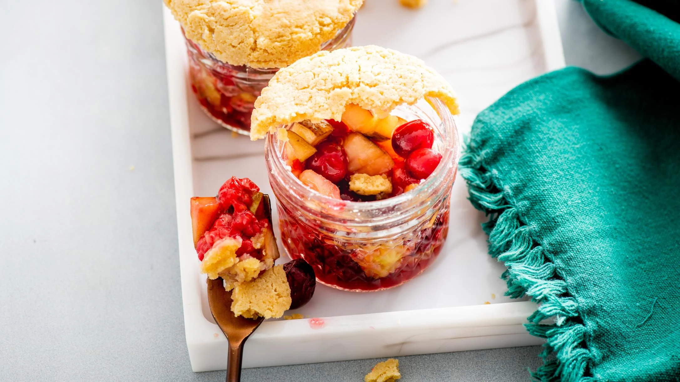 Two glass jars filled with red berry cobbler topped with golden pie crust on a marble tray with a teal napkin.