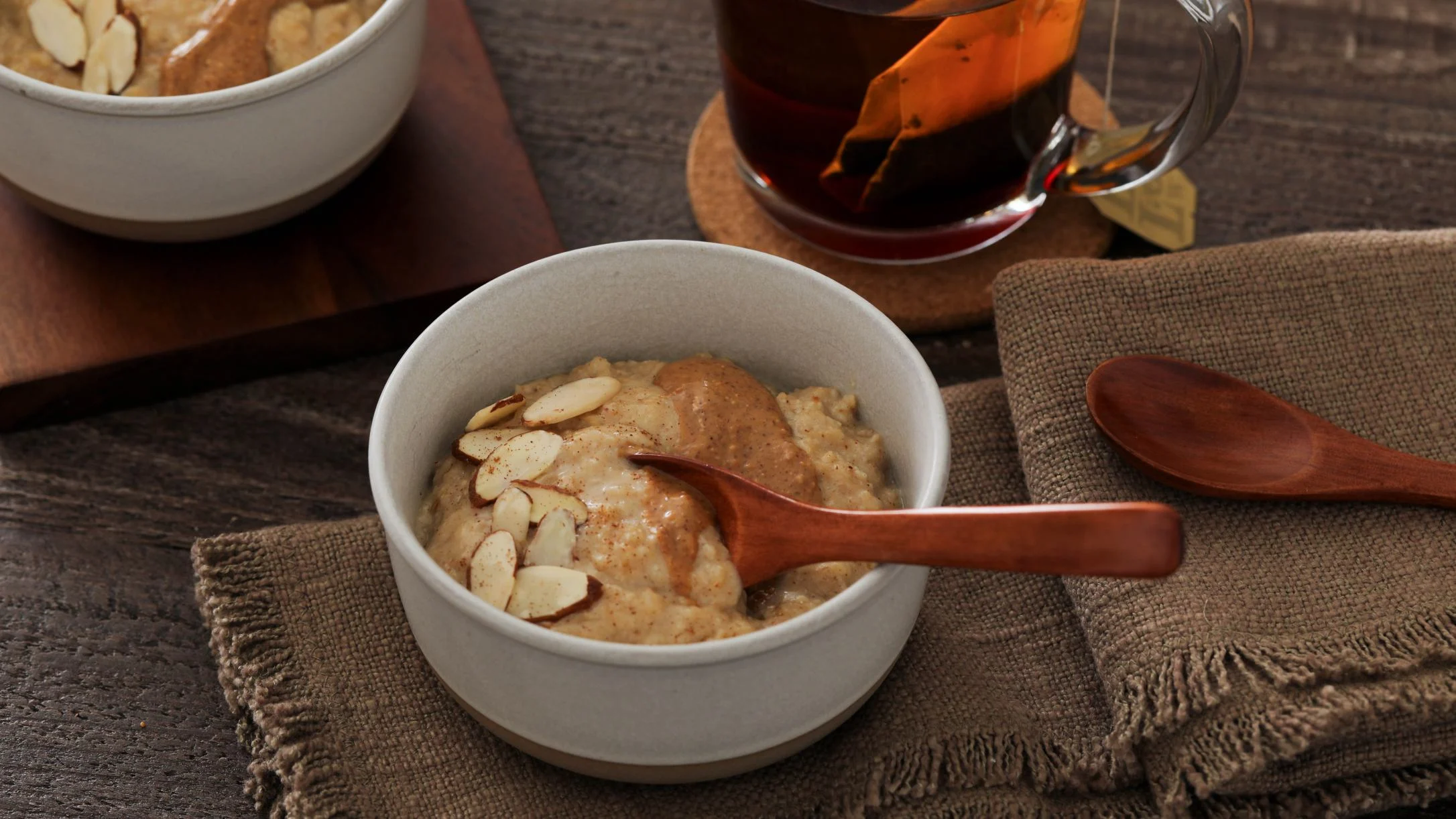 Bowl of oatmeal topped with sliced almonds and cinnamon, wooden spoon, cup of tea with teabag on rustic cloth.