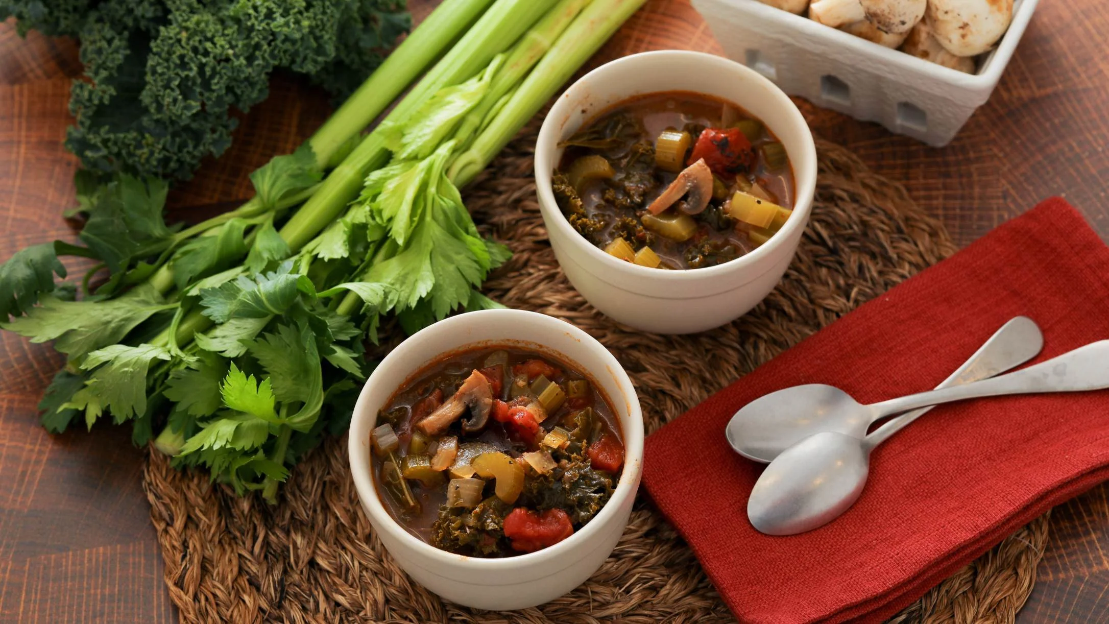Two white bowls of hearty vegetable soup with kale, mushrooms, and tomatoes, beside fresh celery, herbs, and spoons.