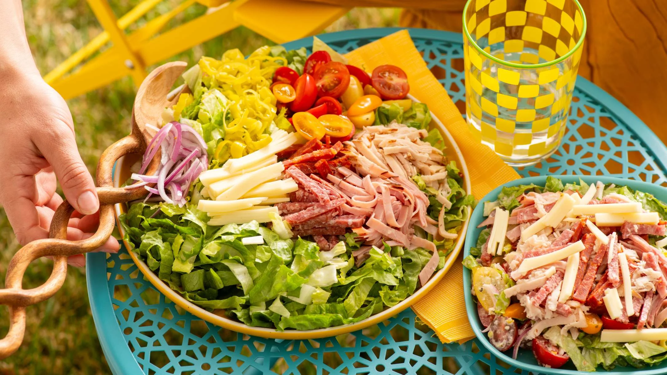 A colorful chopped salad with meats, cheese, tomatoes, and onions in a large bowl on a teal outdoor table.