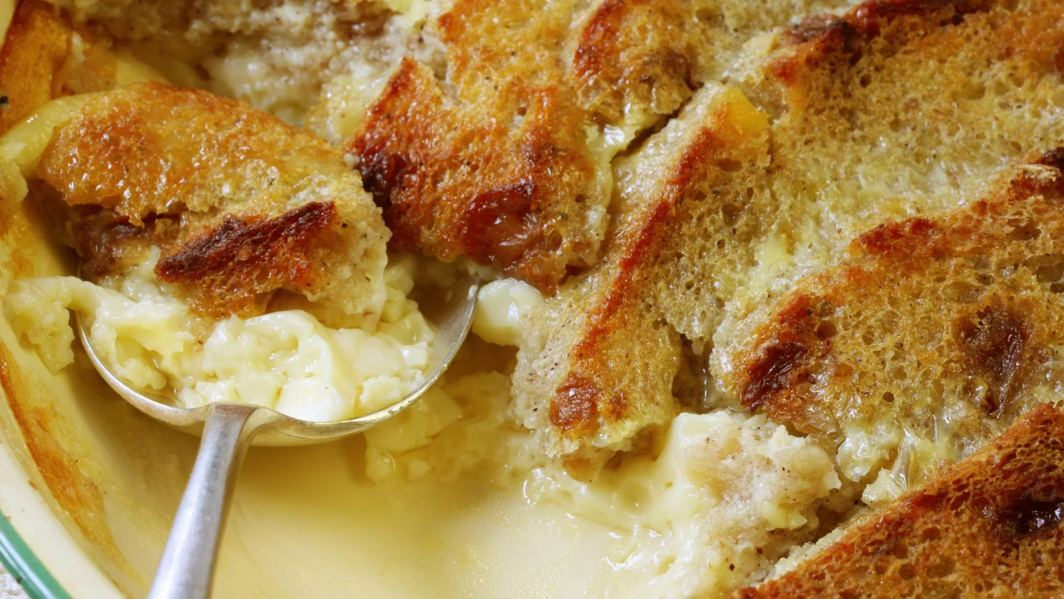 A close-up of a golden bread pudding in a yellow dish, with a spoon scooping out a creamy, custard-filled portion.