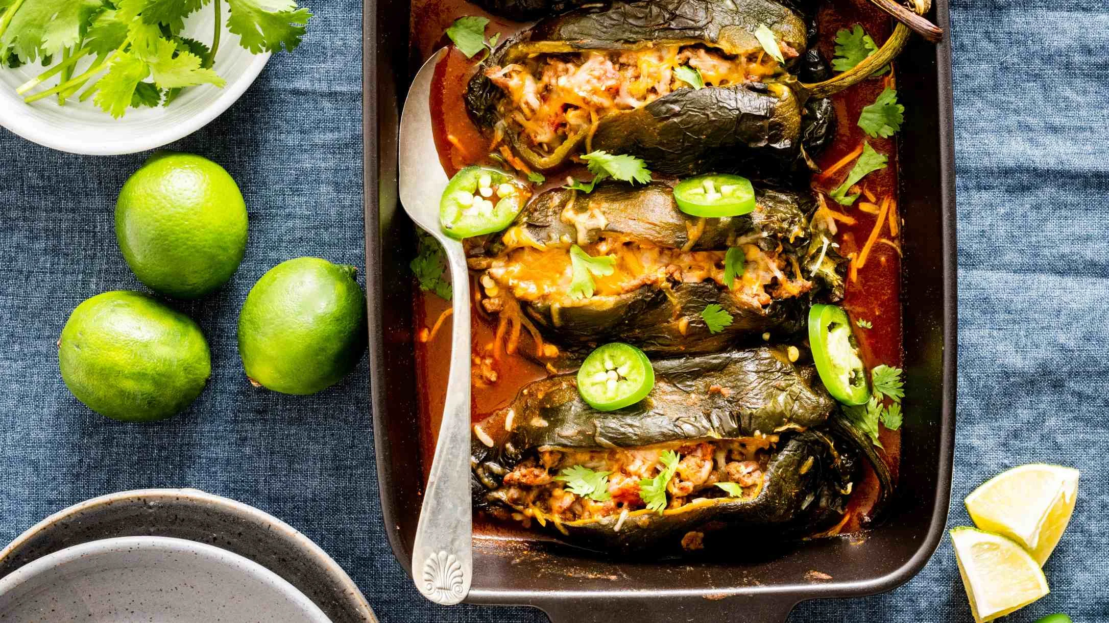 Stuffed poblano peppers in red sauce garnished with jalapeño slices and cilantro, served with fresh limes on blue tablecloth.
