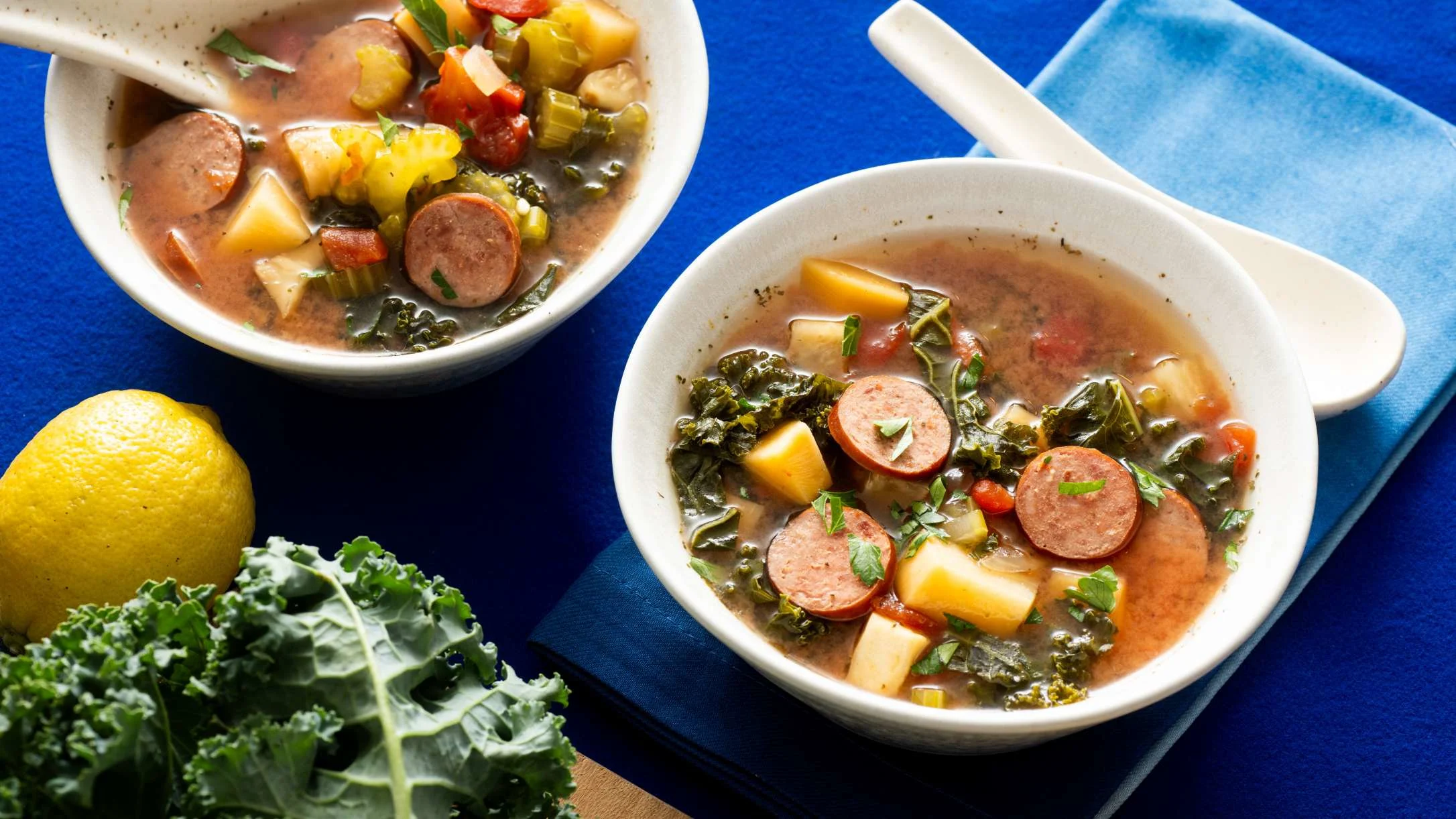Two white bowls of hearty vegetable soup with sliced sausage, potatoes, and kale on a blue background with fresh lemon.