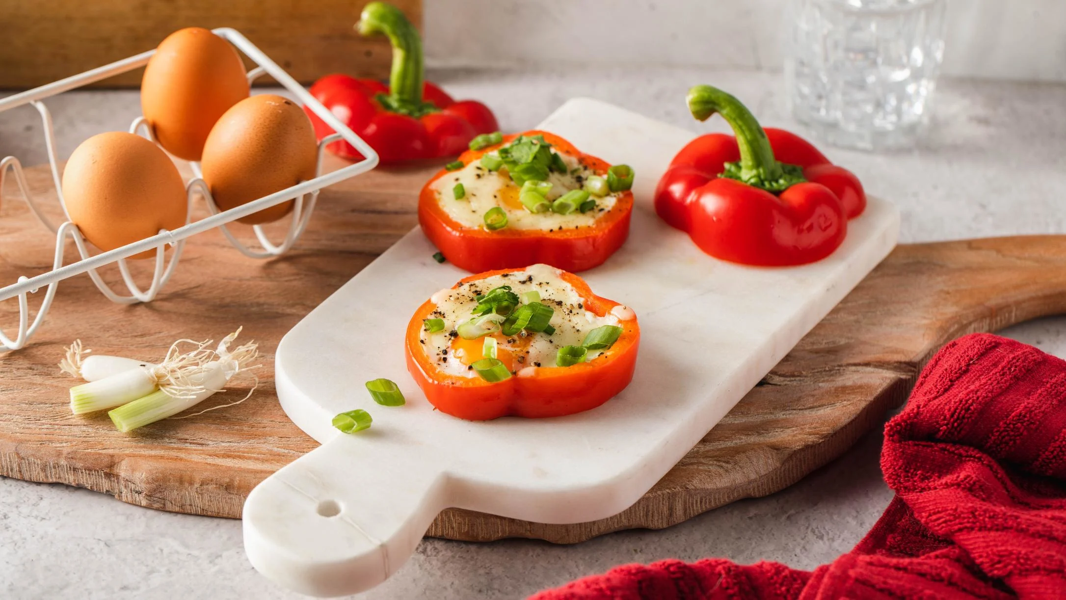 Eggs baked in red bell pepper rings topped with green onions, served on a marble board with whole peppers and eggs nearby.