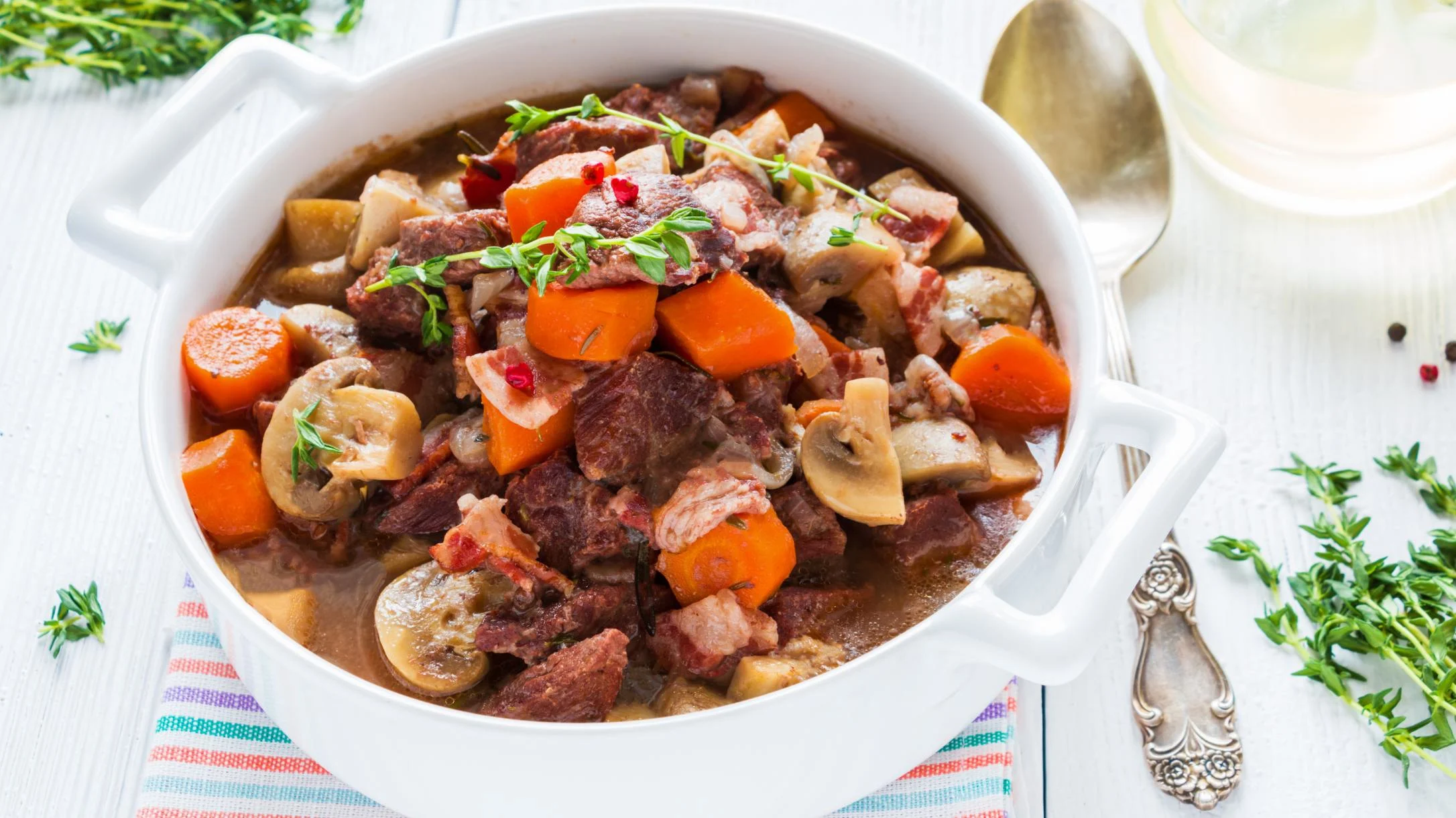 A white casserole dish filled with hearty beef stew featuring carrots, mushrooms, and fresh thyme, served on a white table.