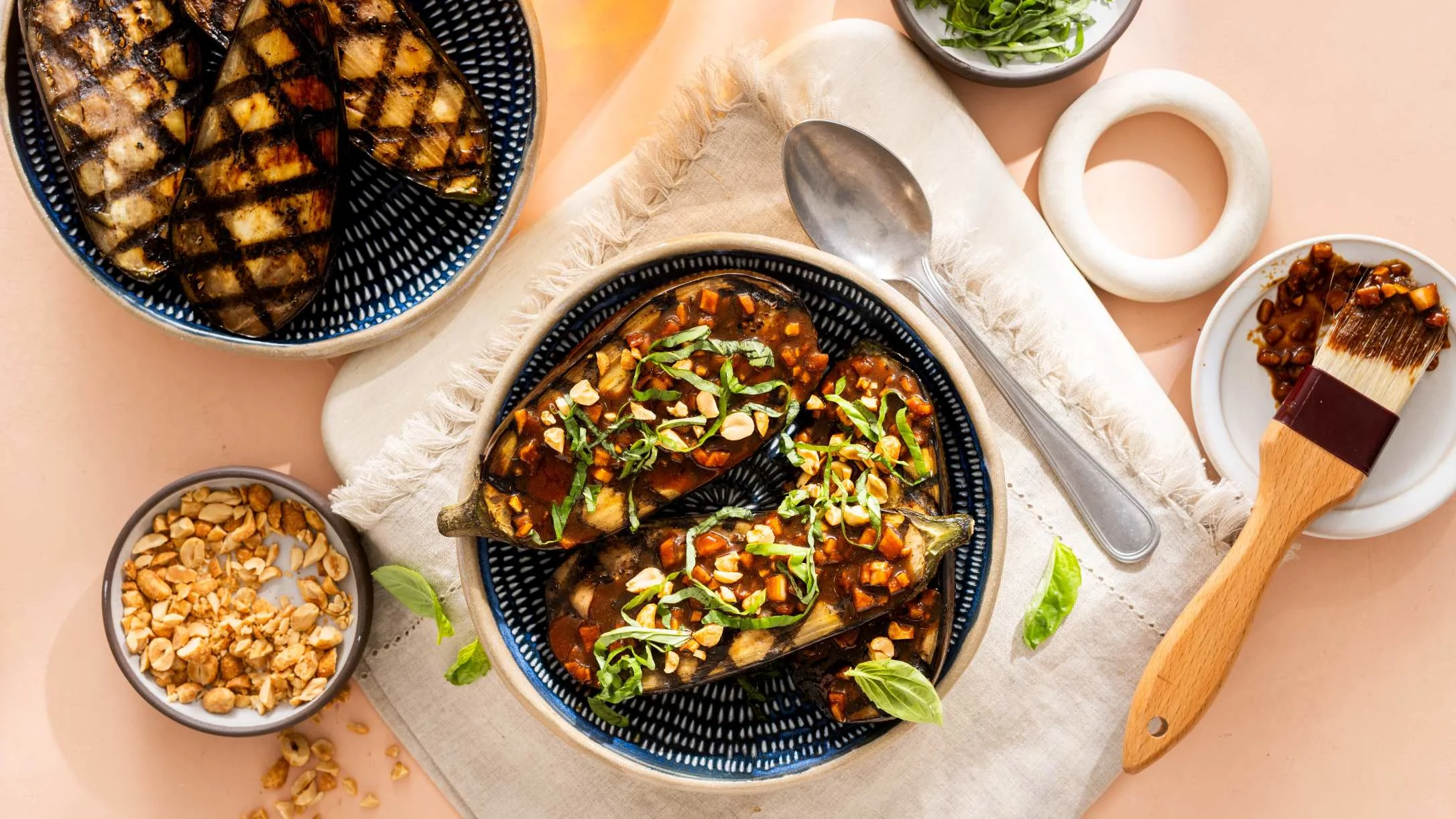 Grilled eggplant halves topped with savory sauce, peanuts, and fresh basil in a blue bowl, surrounded by ingredients.