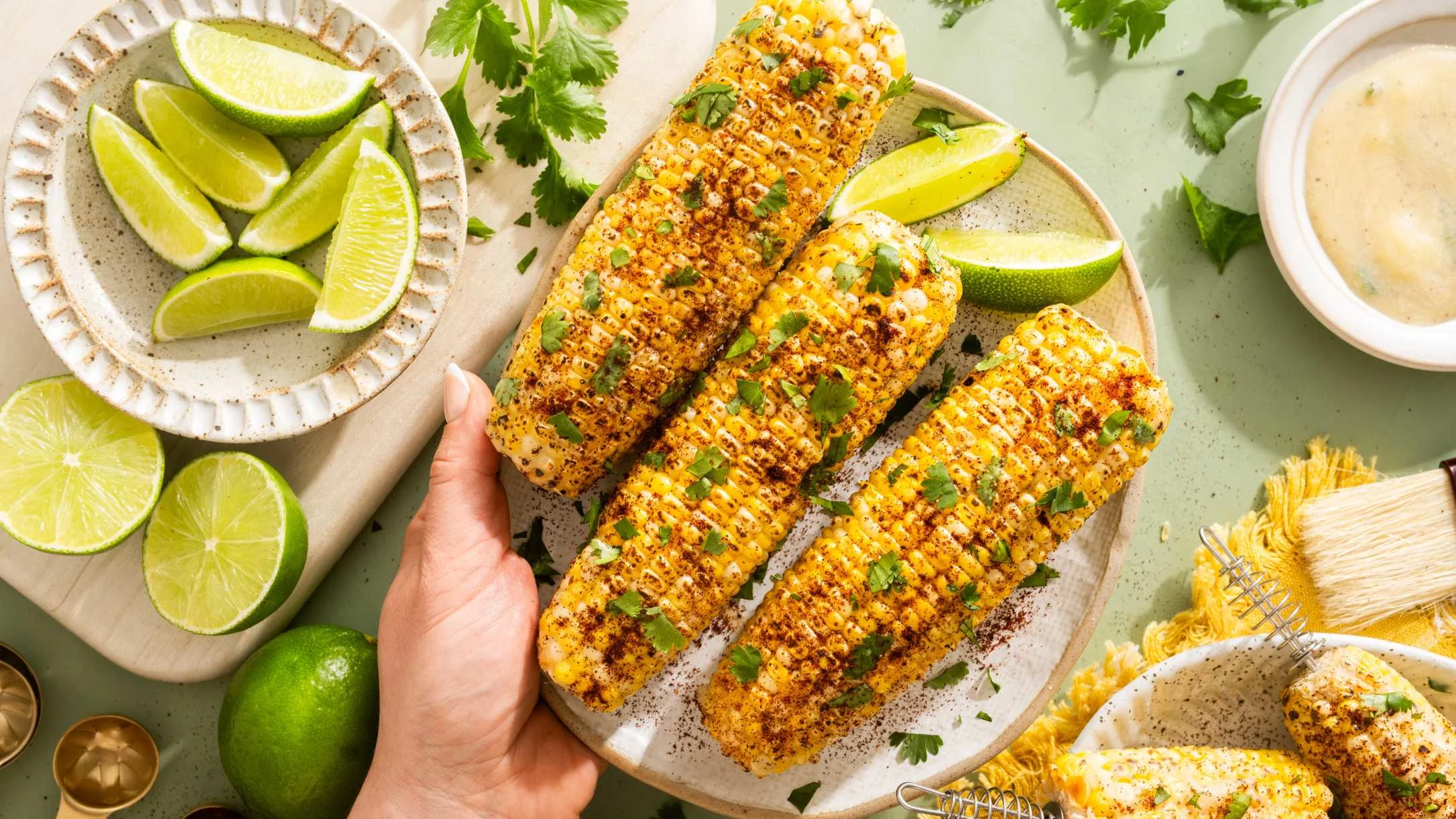 Grilled corn on the cob with spices and cilantro on a ceramic plate, served with lime wedges and dipping sauce.