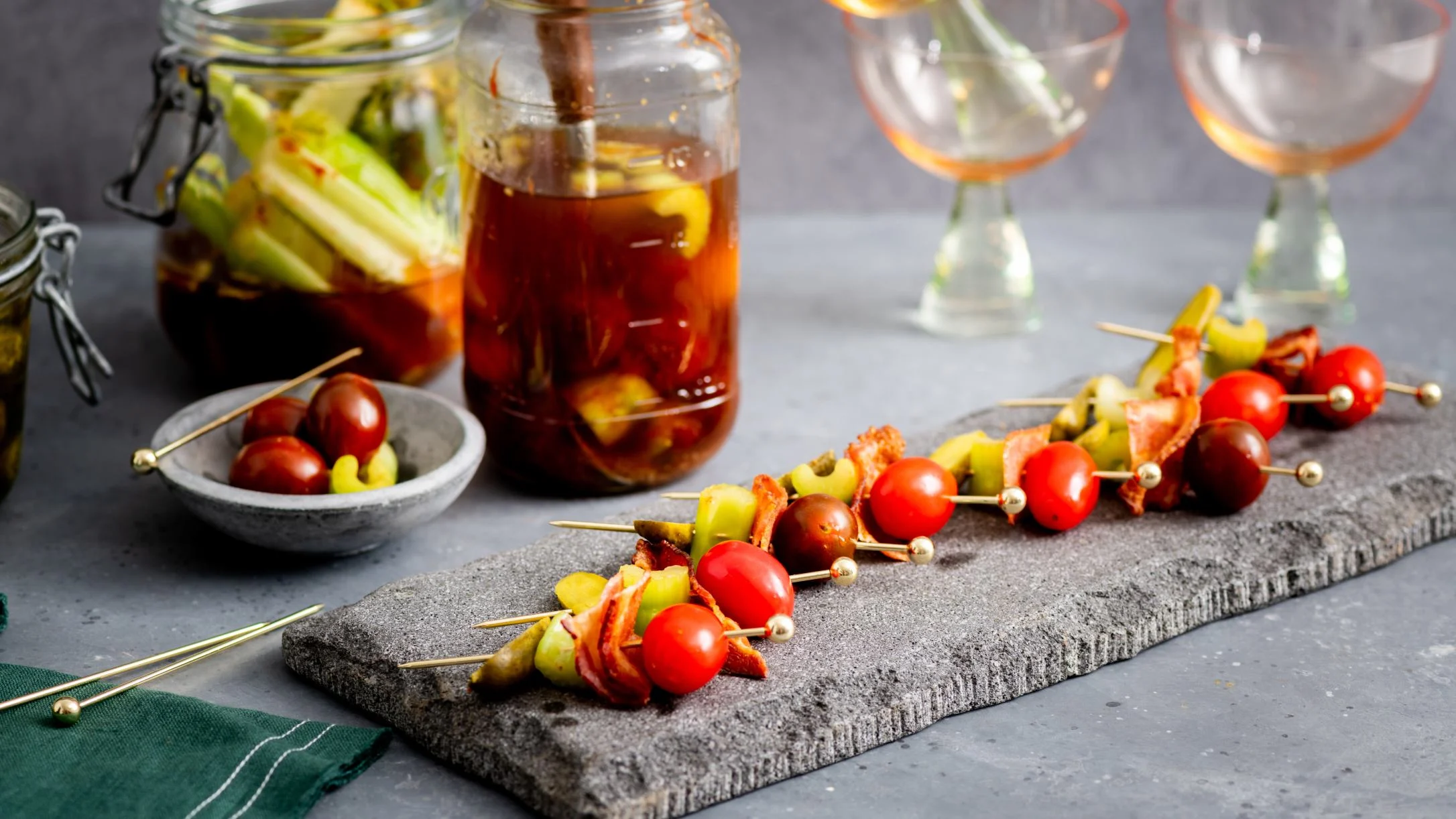 Appetizer skewers with cherry tomatoes and olives on stone board, with pickled vegetables in jars and wine glasses.