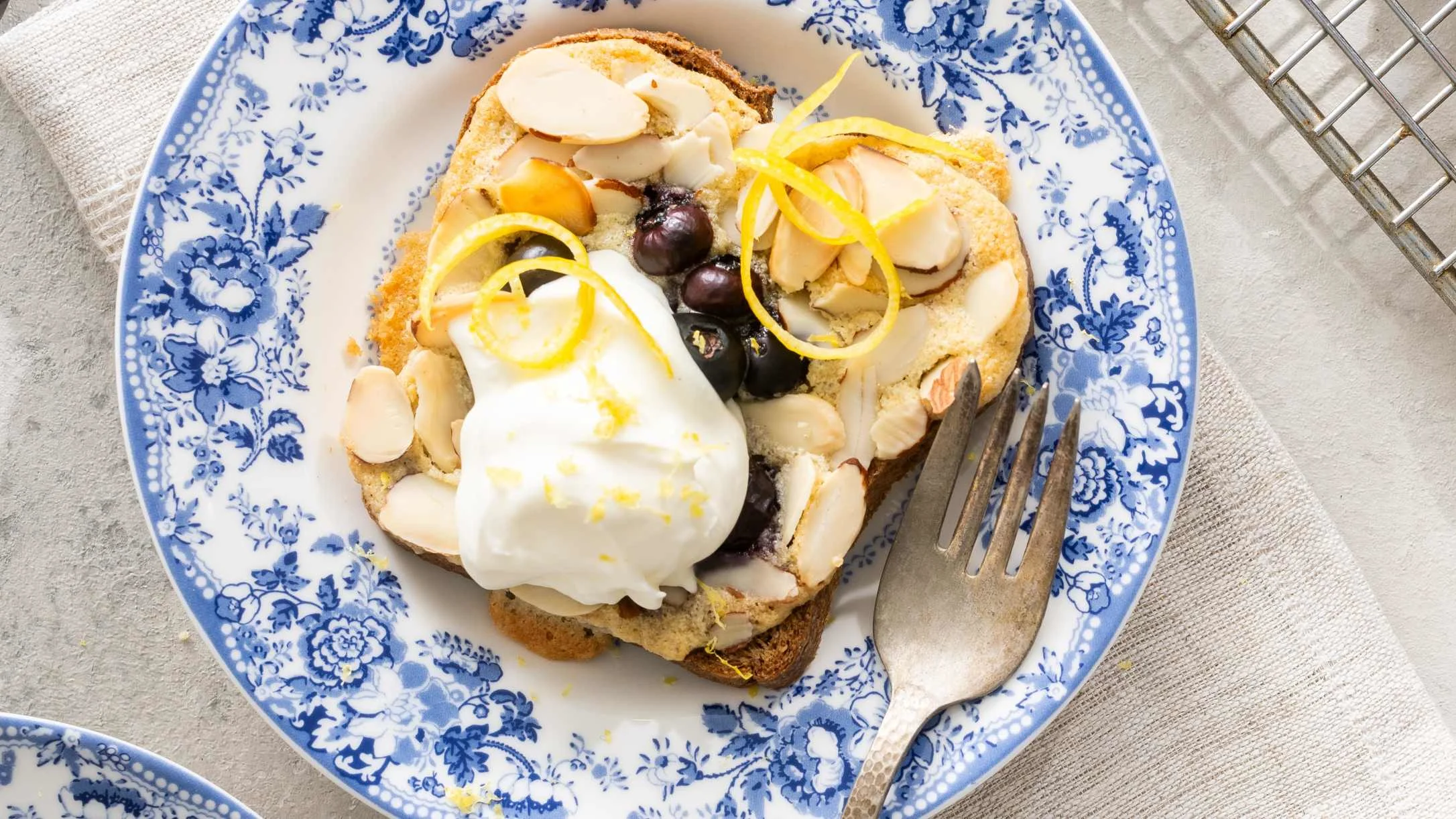 Toast topped with whipped cream, blueberries, sliced almonds, and lemon zest on a blue floral plate with a fork.