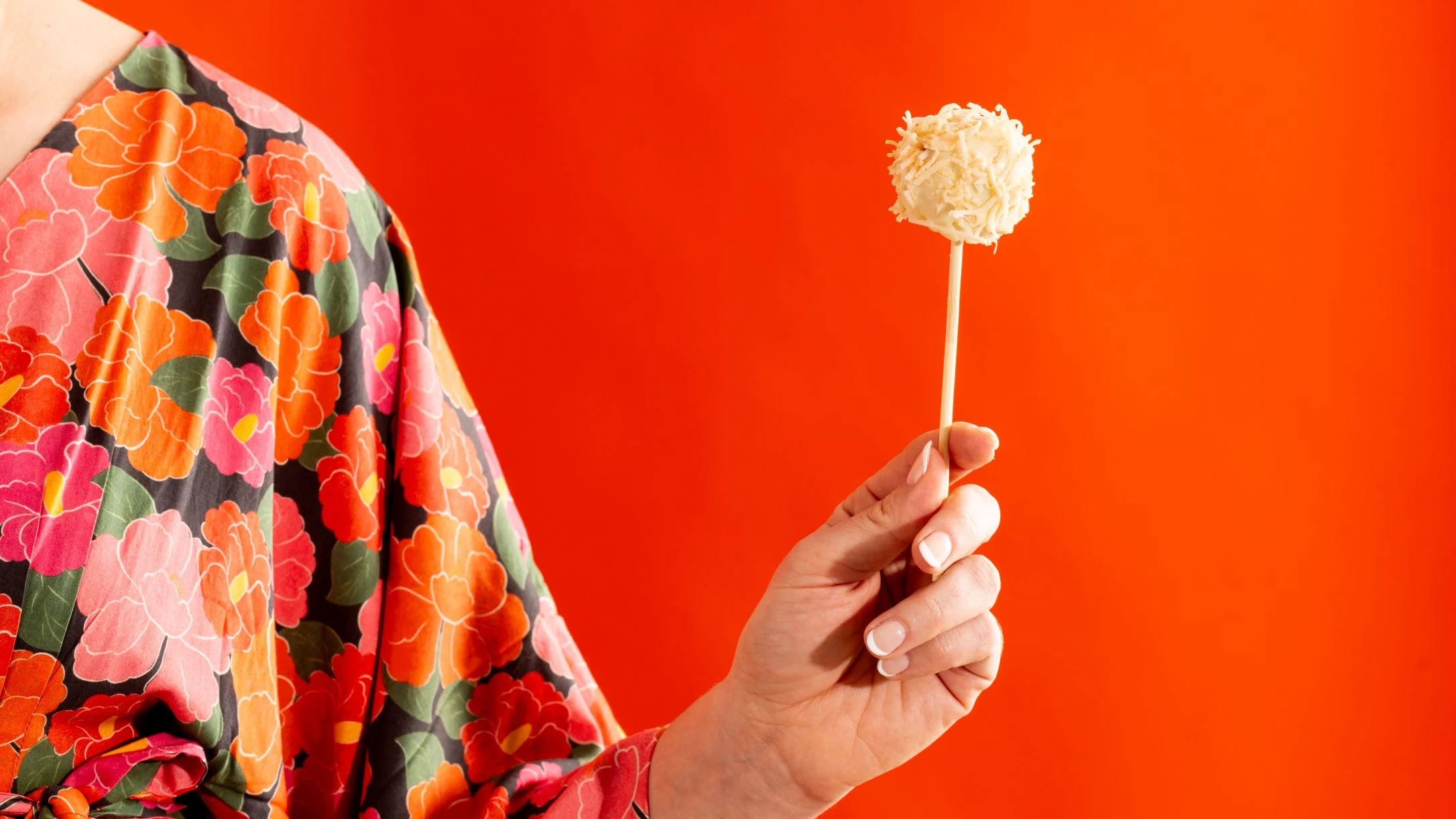 Hand in floral patterned clothing holding a coconut cake pop against a bright orange background.