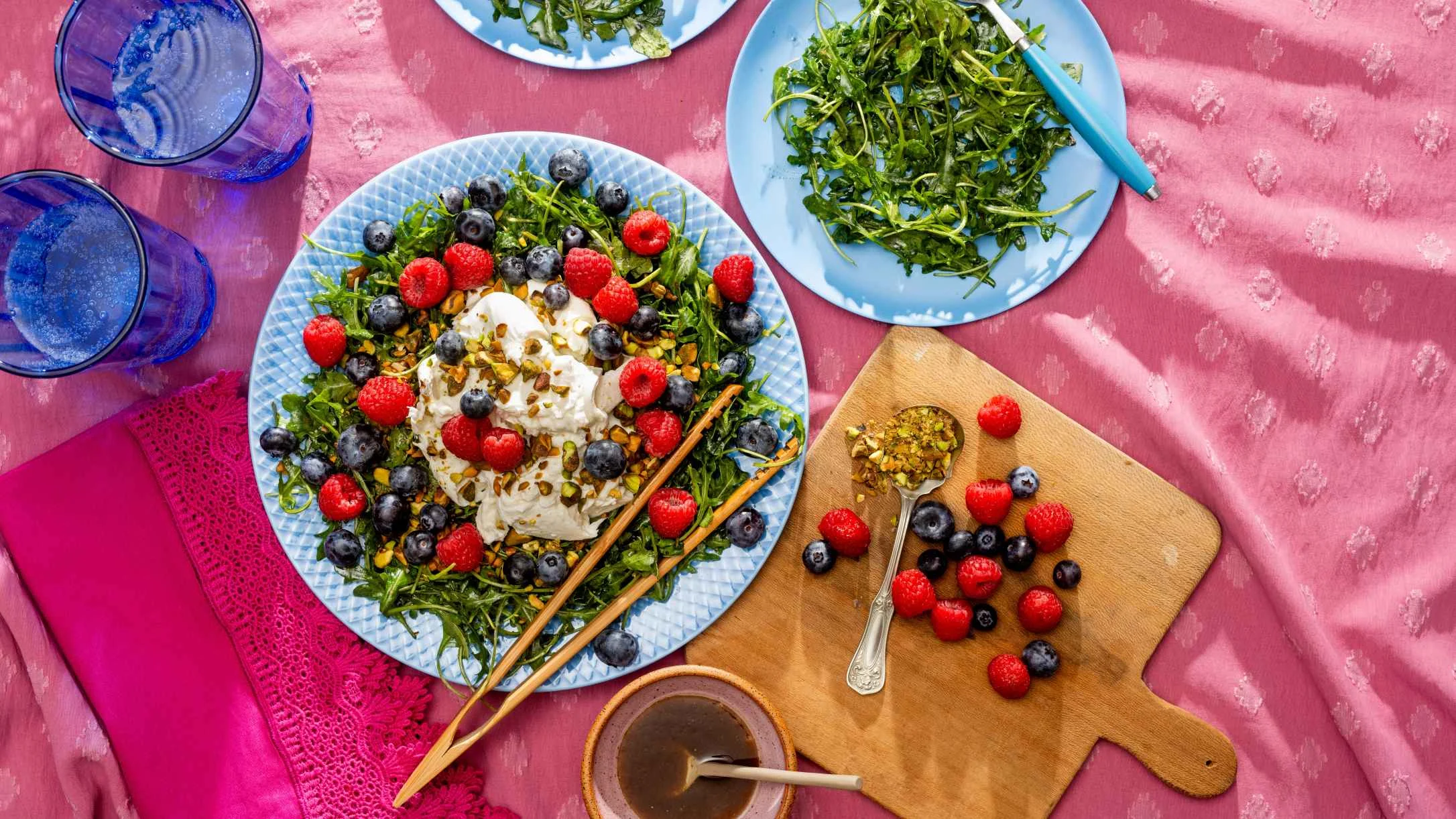 Fresh salad with burrata, berries and greens on blue plate, wooden cutting board with fruits, and blue glasses on pink tablecloth.