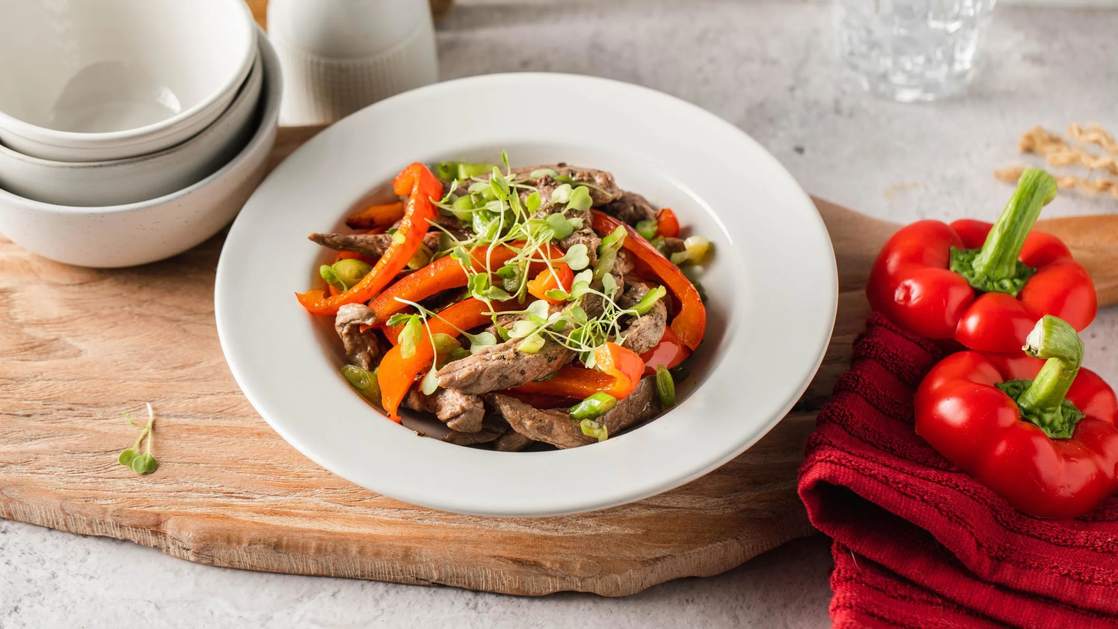 Beef sauté with red bell peppers and microgreens in white bowl, with red bell peppers and stacked bowls nearby.