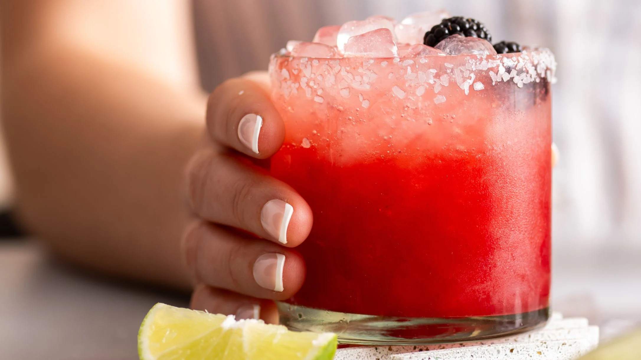 A hand holding a red cocktail in a salt-rimmed glass with ice and blackberries, alongside a lime wedge.