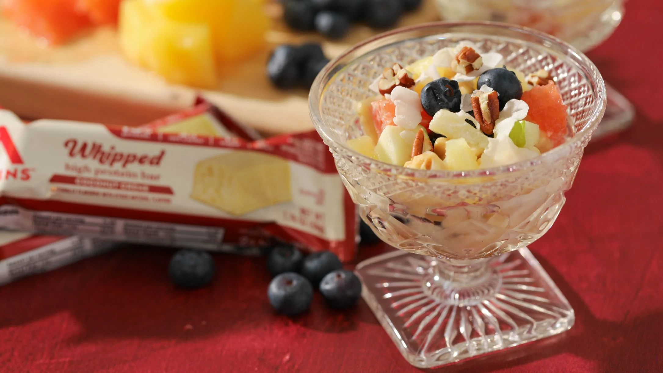 A crystal dessert cup filled with fruit salad, blueberries, and pecans, beside an Atkins Whipped high-protein bar package on a red surface.