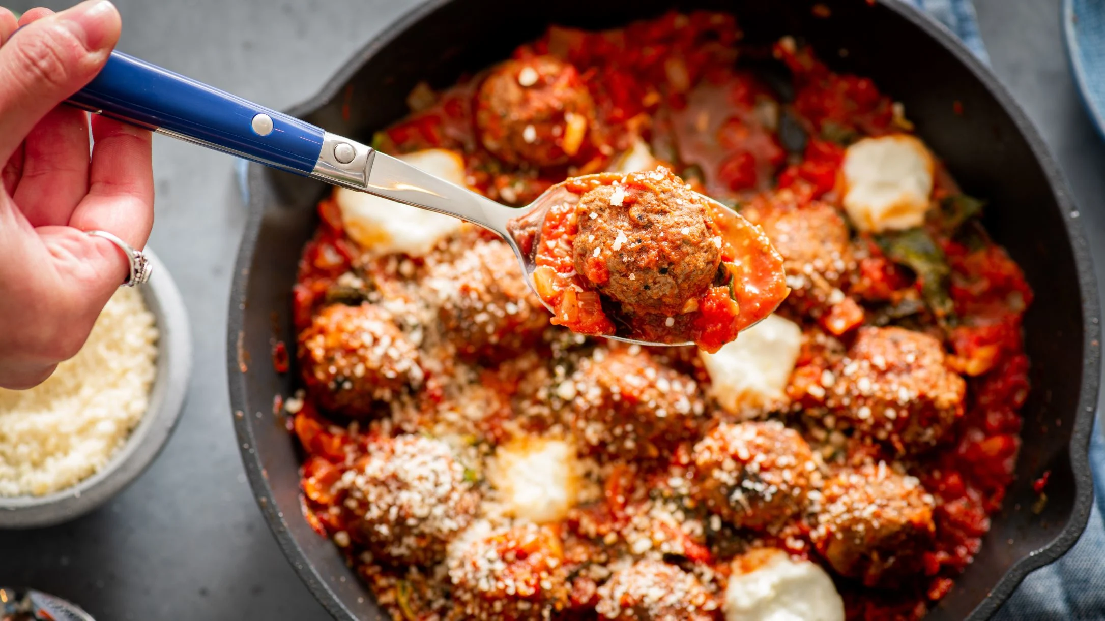 A hand lifts a meatball with a blue-handled spoon from a cast iron skillet filled with tomato sauce, ricotta cheese, and Parmesan.