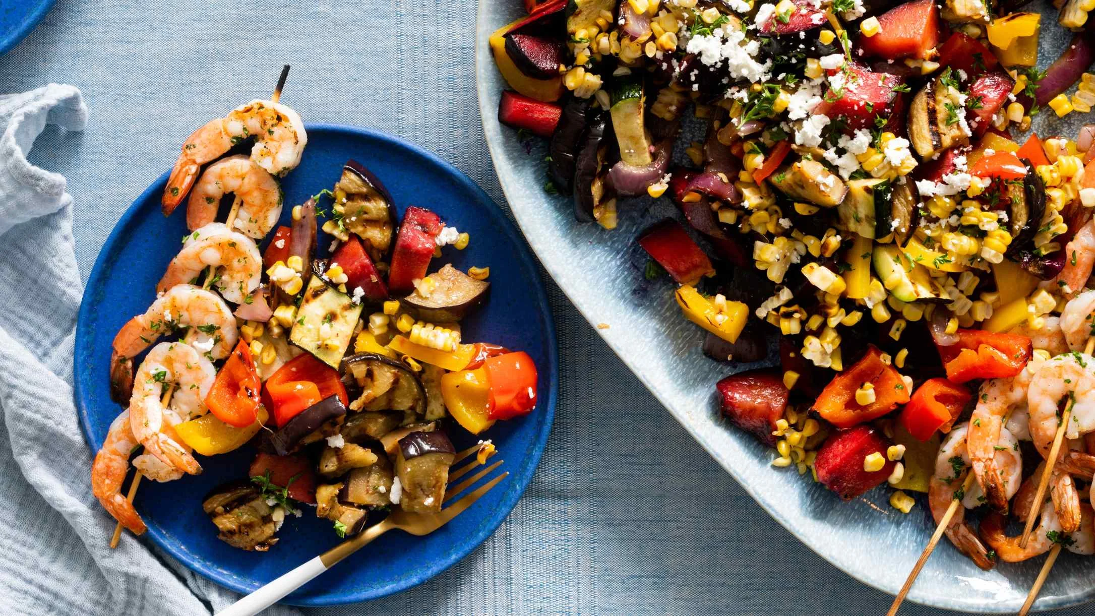 Grilled shrimp skewers with colorful roasted vegetables including corn, eggplant, and bell peppers, topped with crumbled feta.