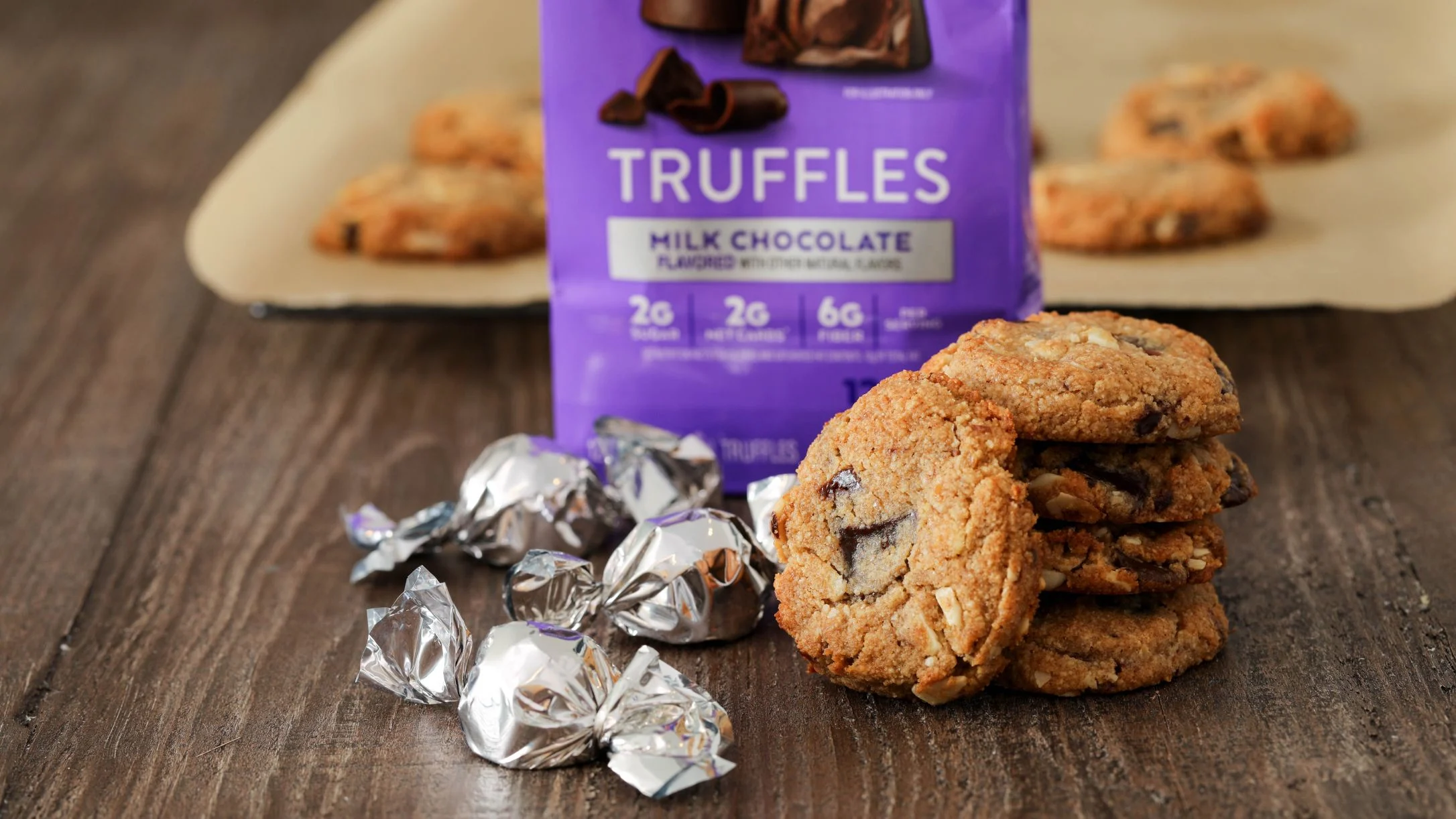 Stacked chocolate chip cookies and silver-wrapped milk chocolate truffles beside a purple Truffles candy bag on a wood surface.