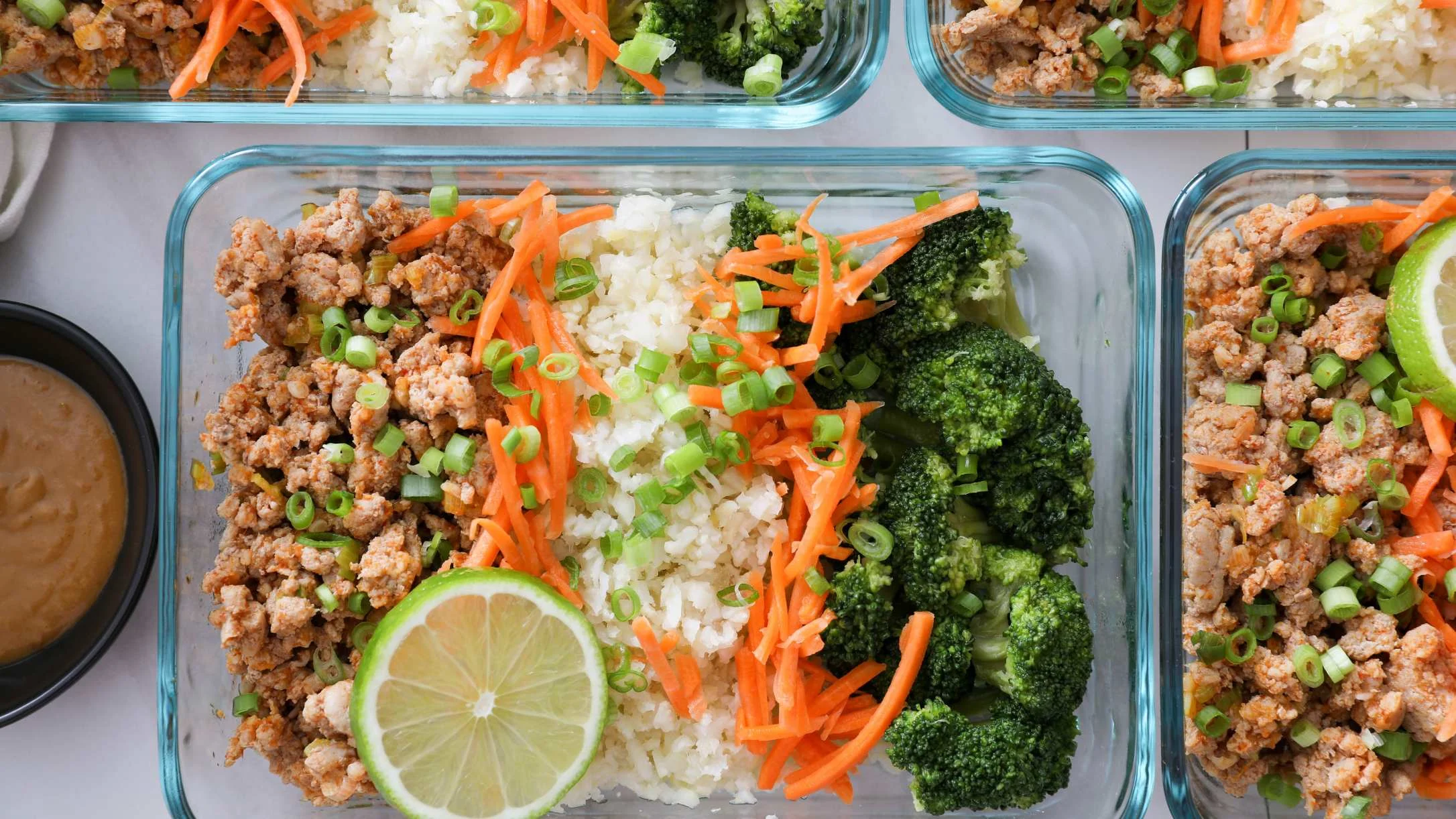 Glass meal prep containers filled with ground meat, cauliflower rice, shredded carrots, broccoli, green onions, and lime.