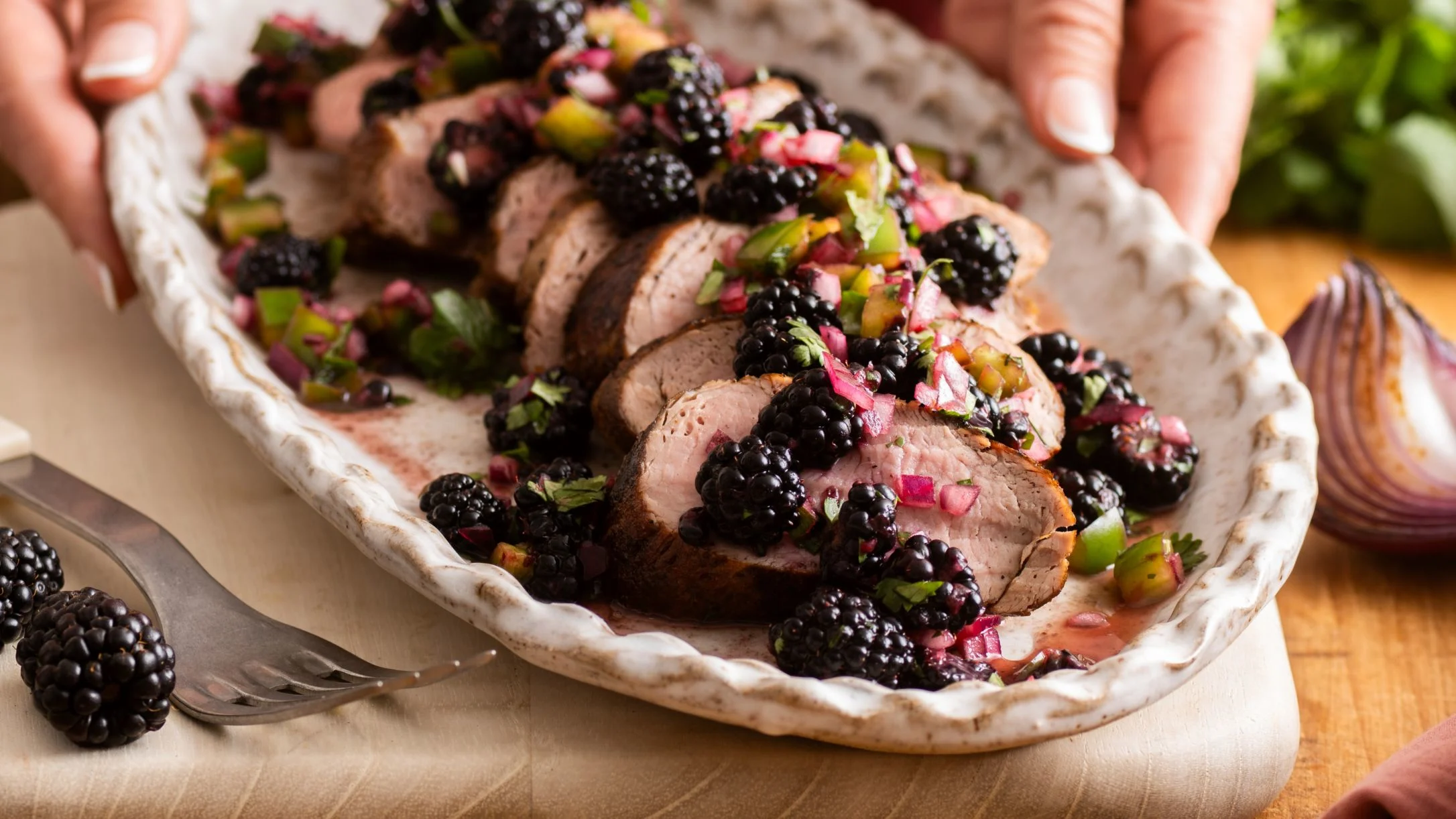 Sliced pork tenderloin topped with blackberry salsa on a rustic white platter, held by two hands, with a fork alongside.