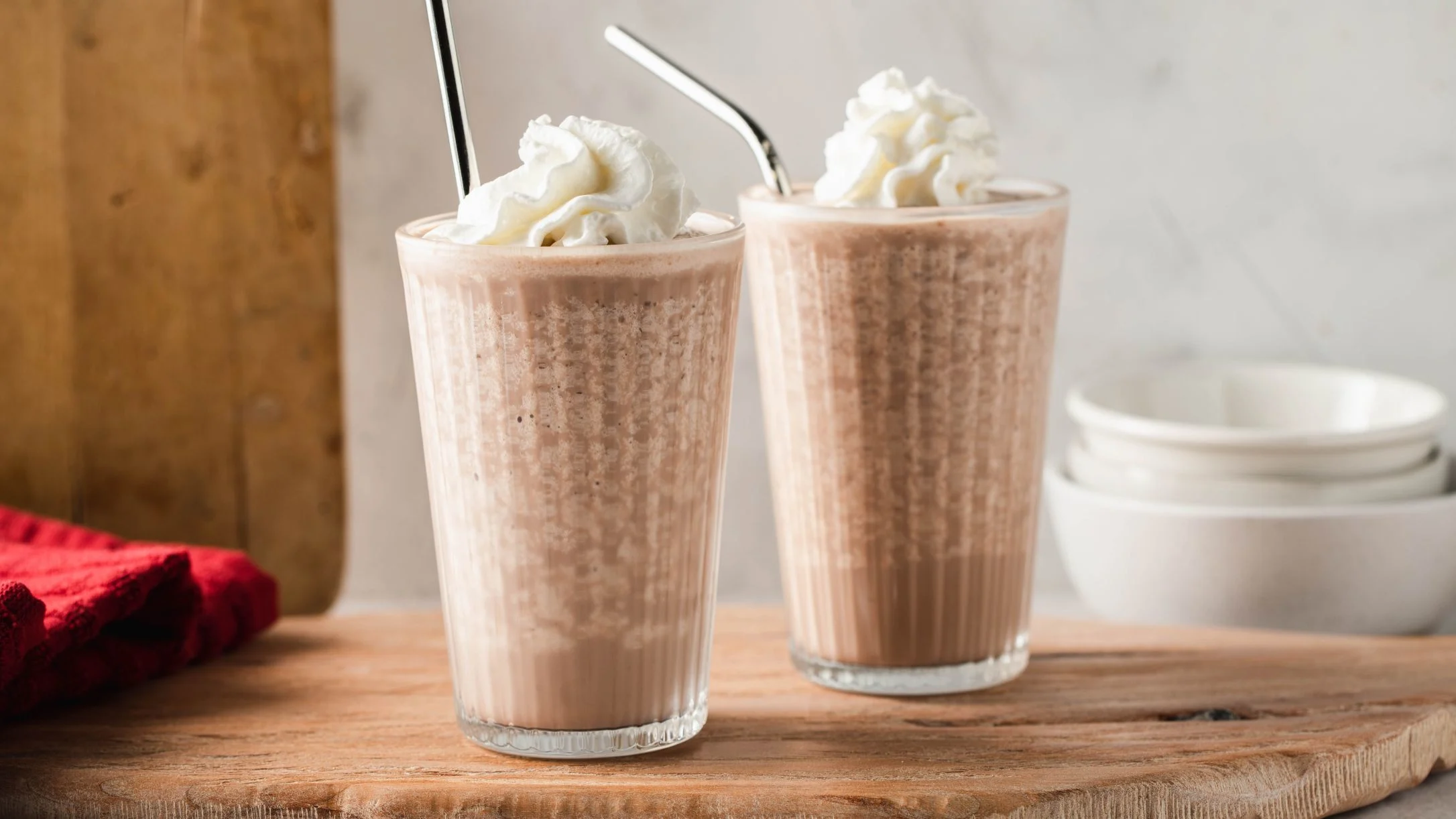 Two chocolate milkshakes topped with whipped cream and metal straws on a wooden board with a red napkin nearby.