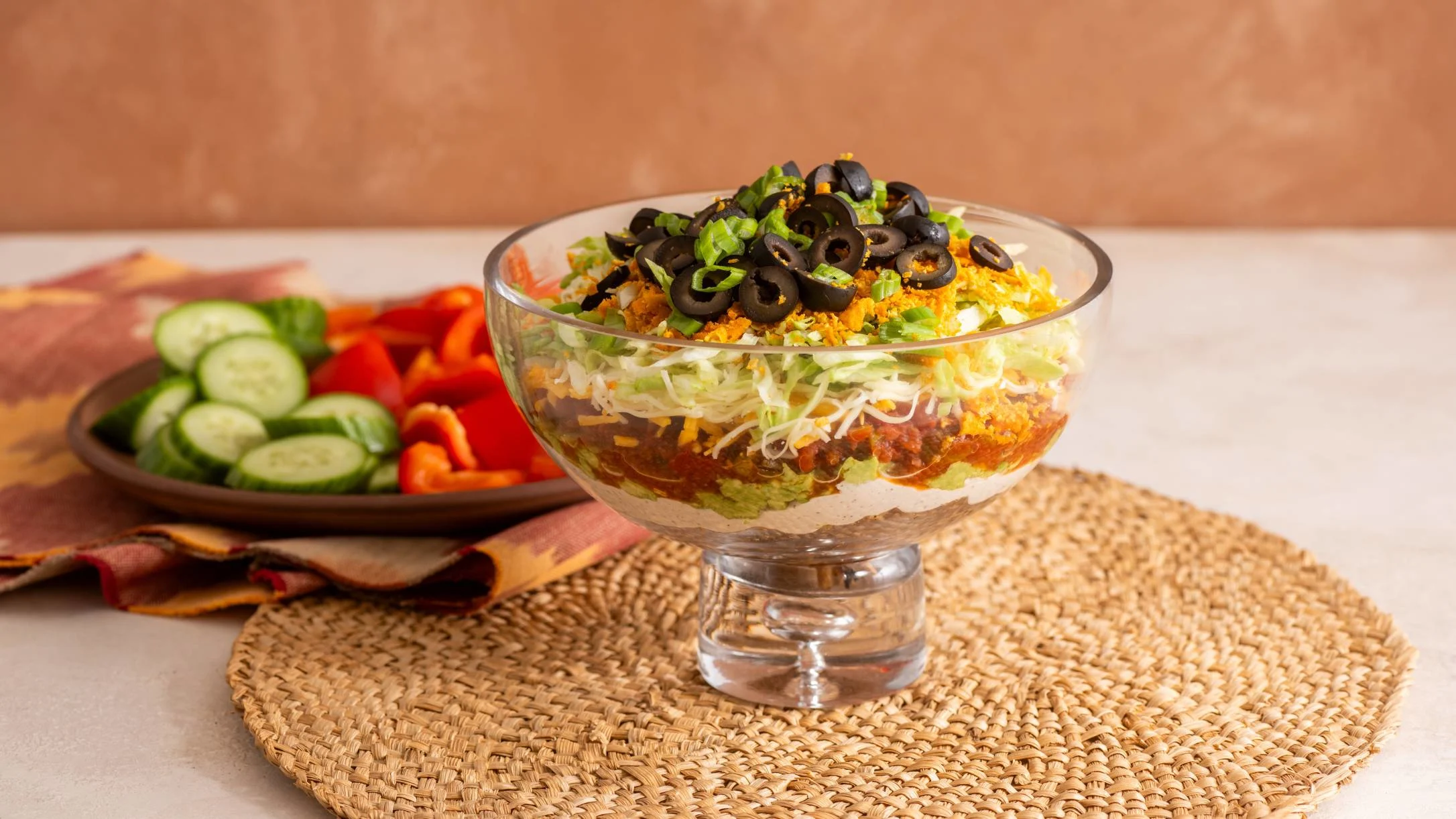 Layered taco dip in glass bowl topped with black olives and green onions, served with cucumber and red pepper slices.