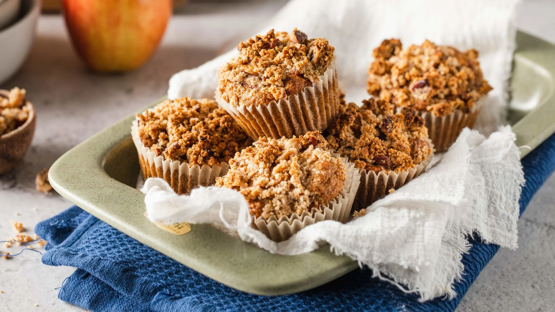 Freshly baked streusel-topped muffins on a green plate with white cloth and blue napkin, with an apple in background.