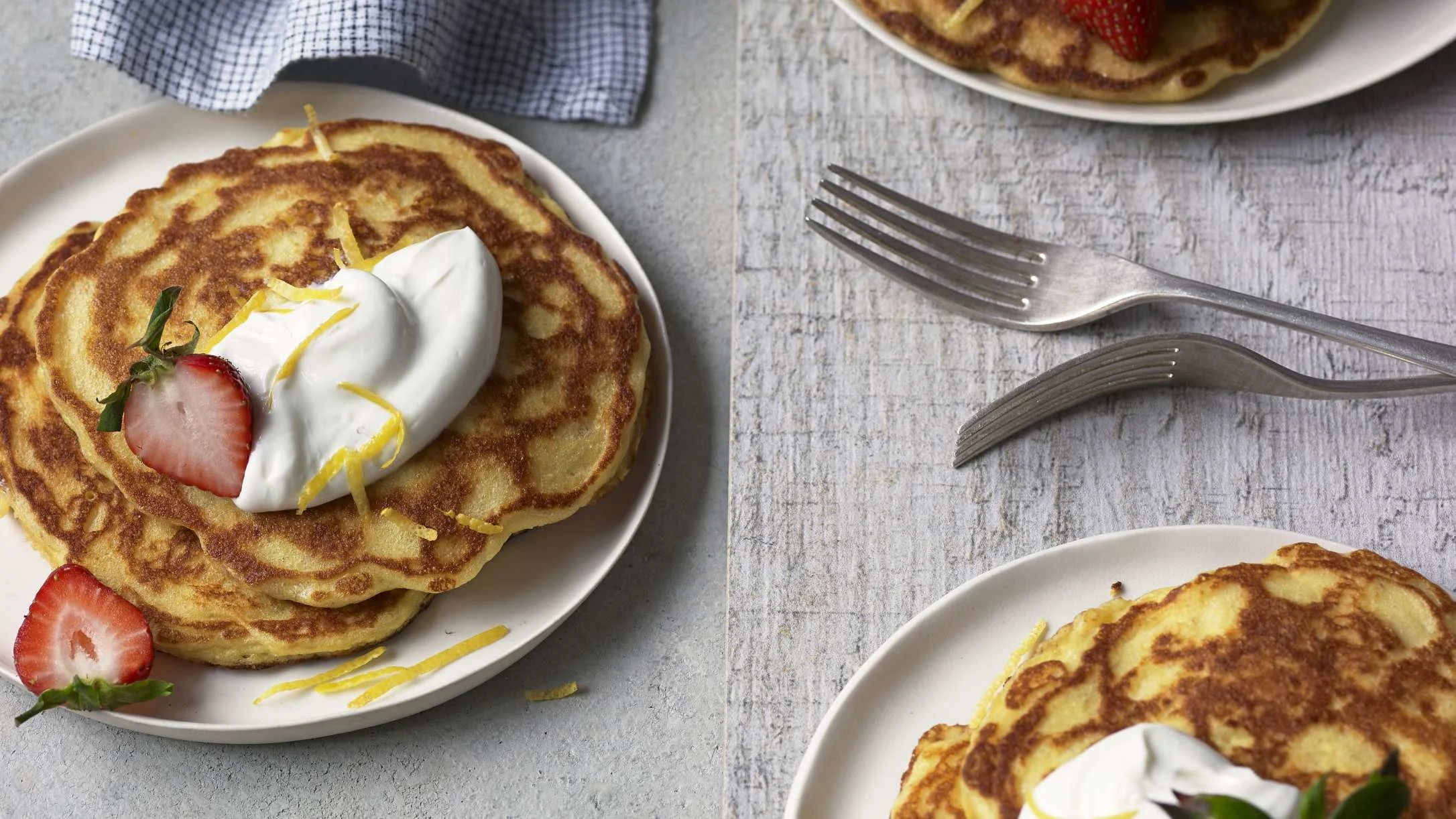 Golden pancakes topped with whipped cream, lemon zest, and fresh strawberries on white plates with forks nearby.