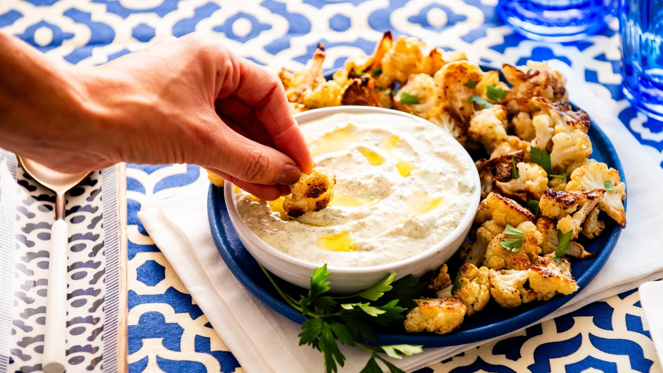 Hand dipping roasted cauliflower into creamy dip on blue plate with patterned tablecloth underneath.