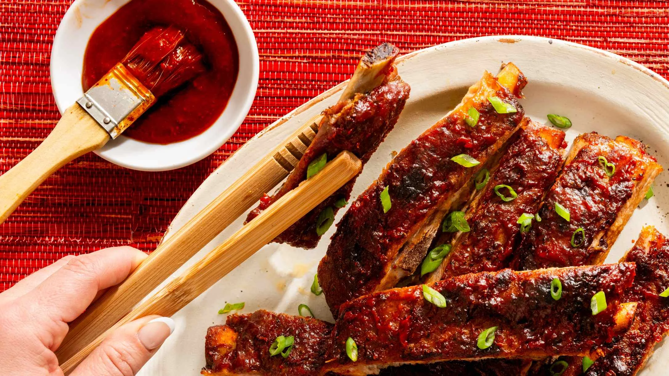 Glazed barbecue ribs garnished with green onions on white plate, with sauce bowl and basting brush on red placemat.