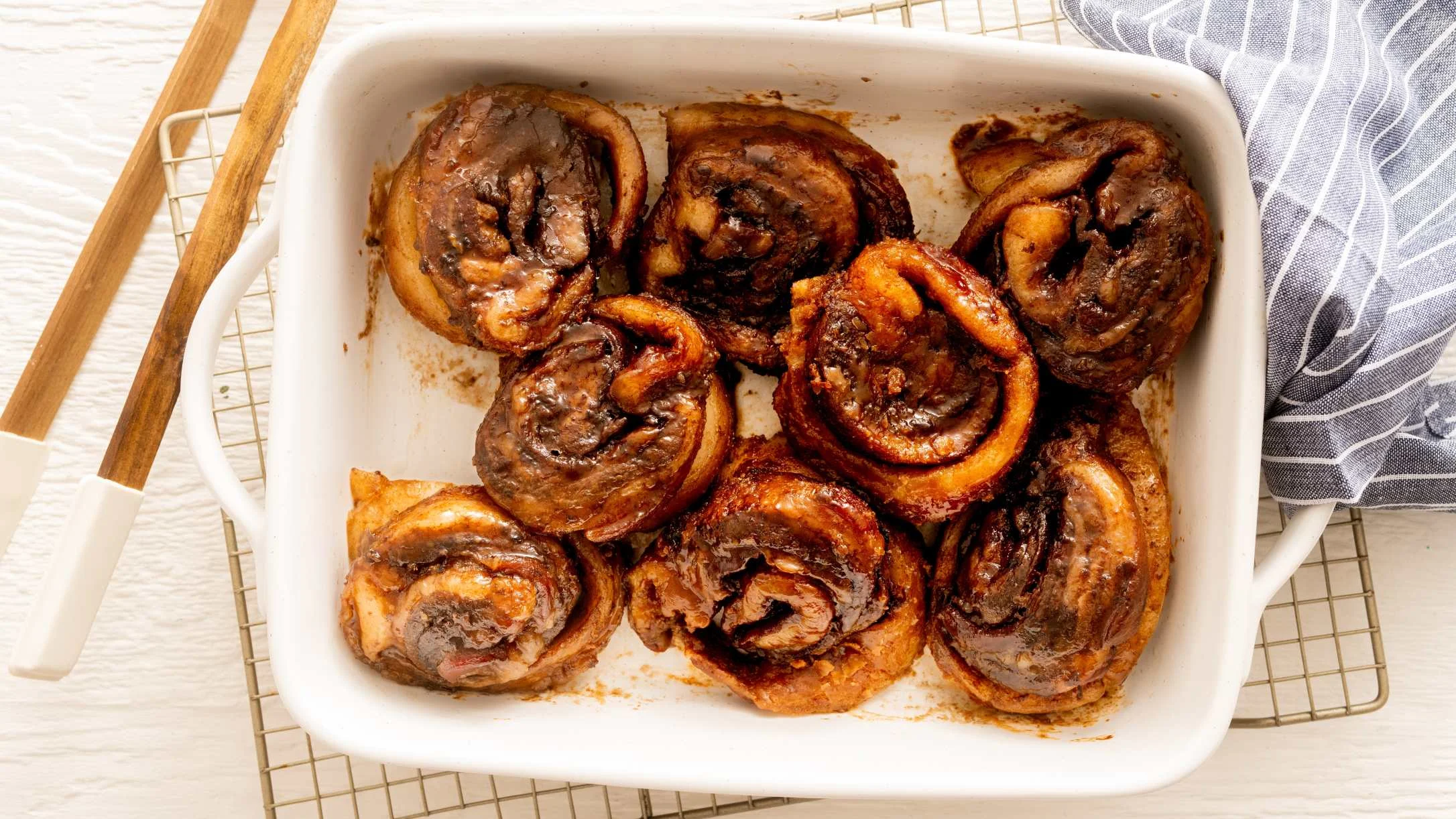 Freshly baked pork belly cinnamon rolls in a white baking dish on a wire rack, with a gray cloth napkin nearby.