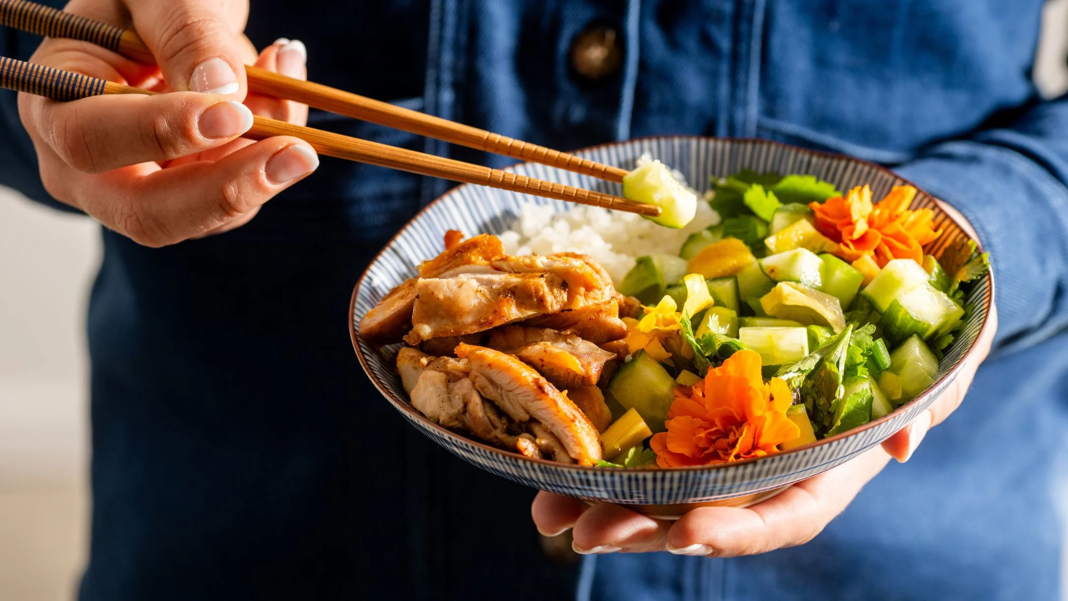 Person holding a colorful rice bowl with chicken, vegetables, and edible flowers while using chopsticks.