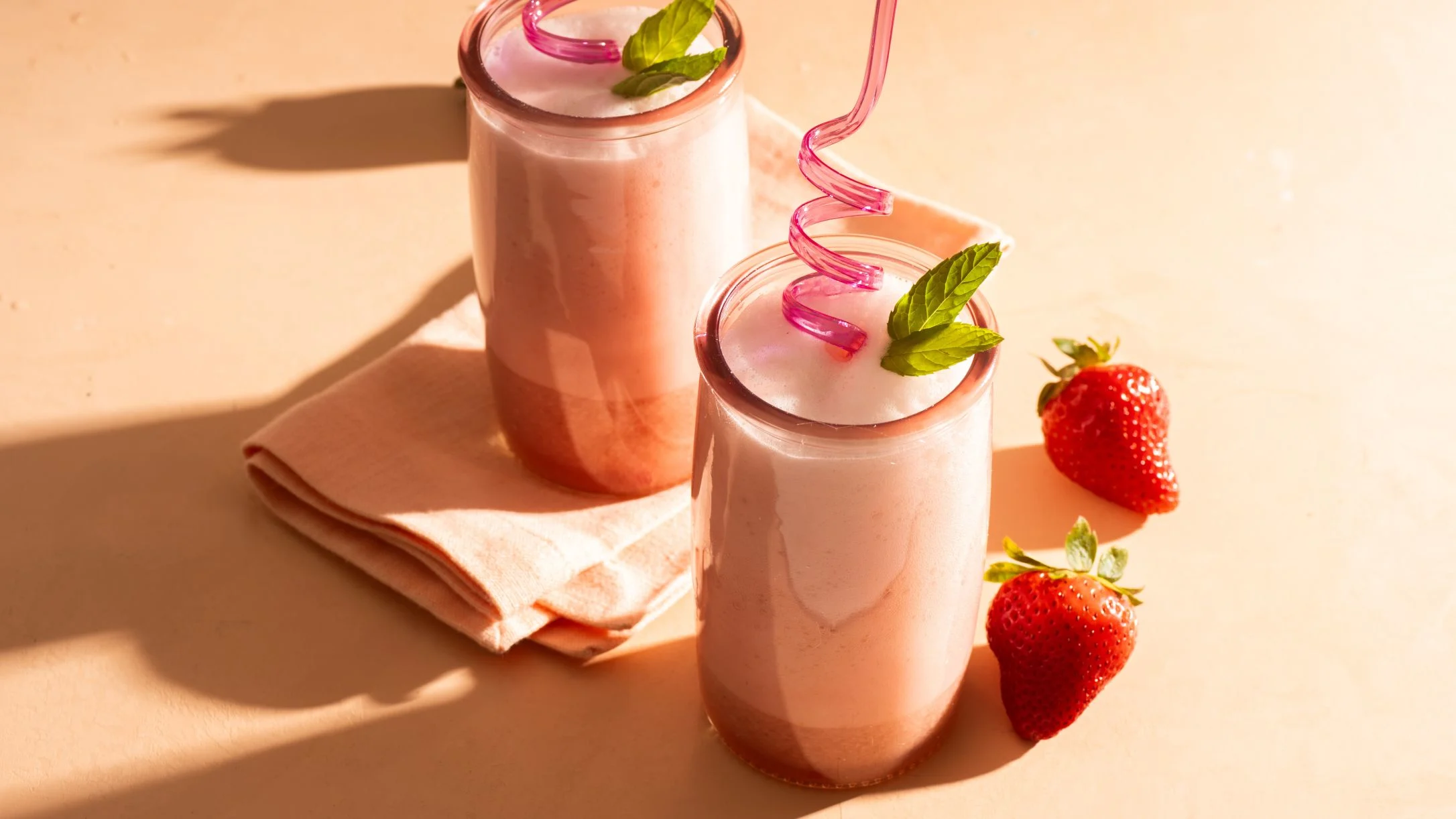 Two pink strawberry smoothies in glass jars with curly straws, mint leaves, and fresh strawberries on peach background.