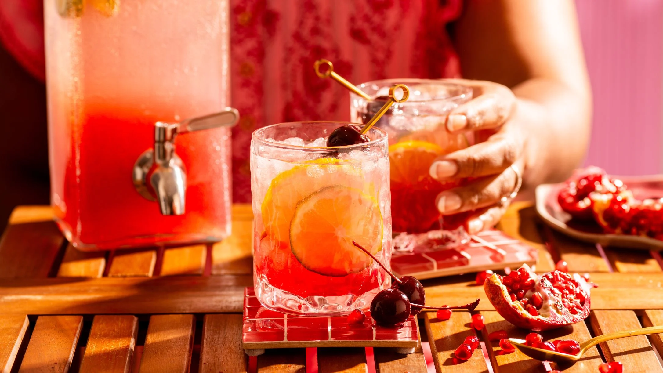 Red cocktail with lemon slices, cherries, and ice on a wooden table beside a drink dispenser and pomegranate.