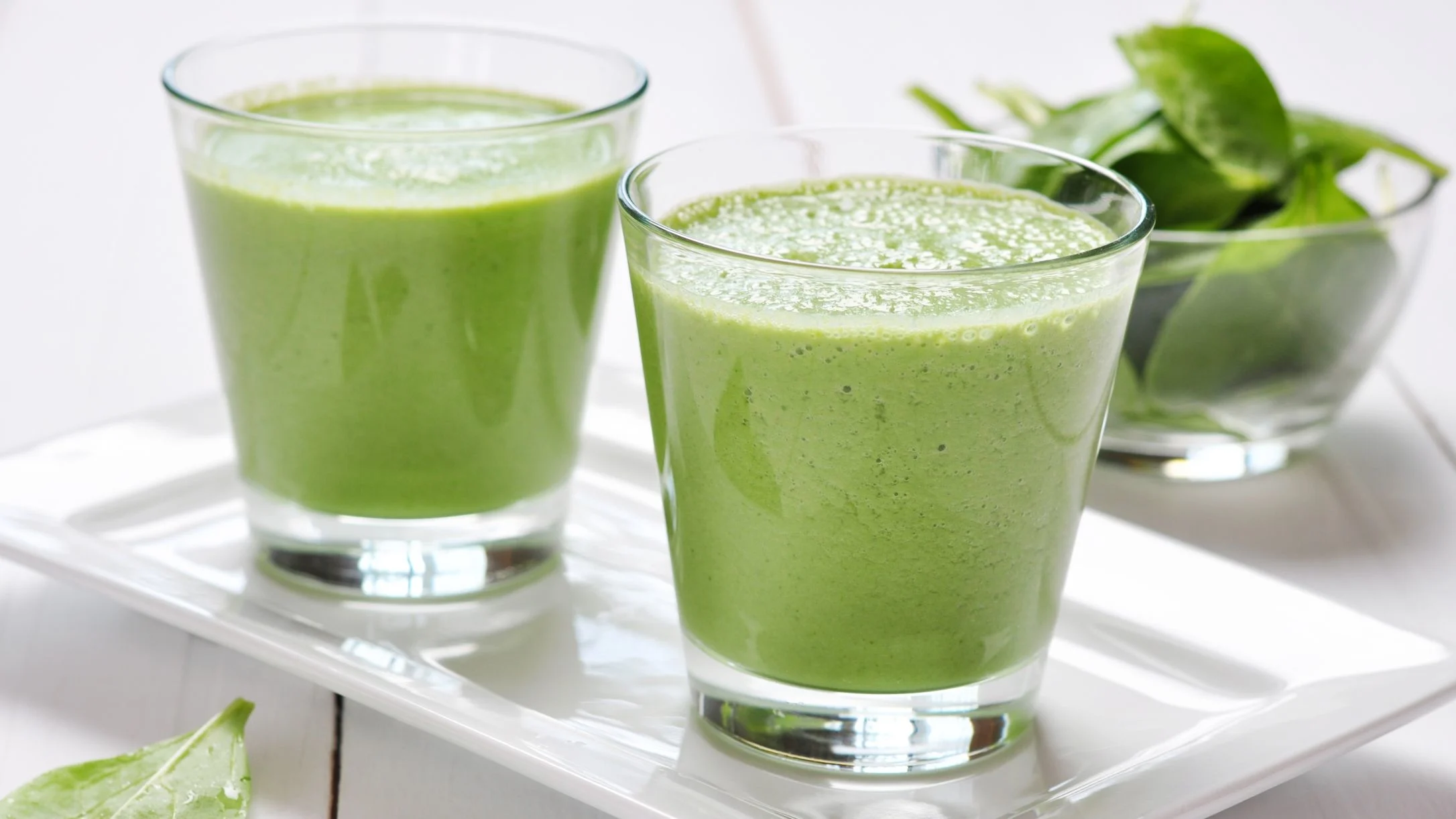 Two glasses of green smoothie on a white tray, with a bowl of fresh spinach leaves in the background.