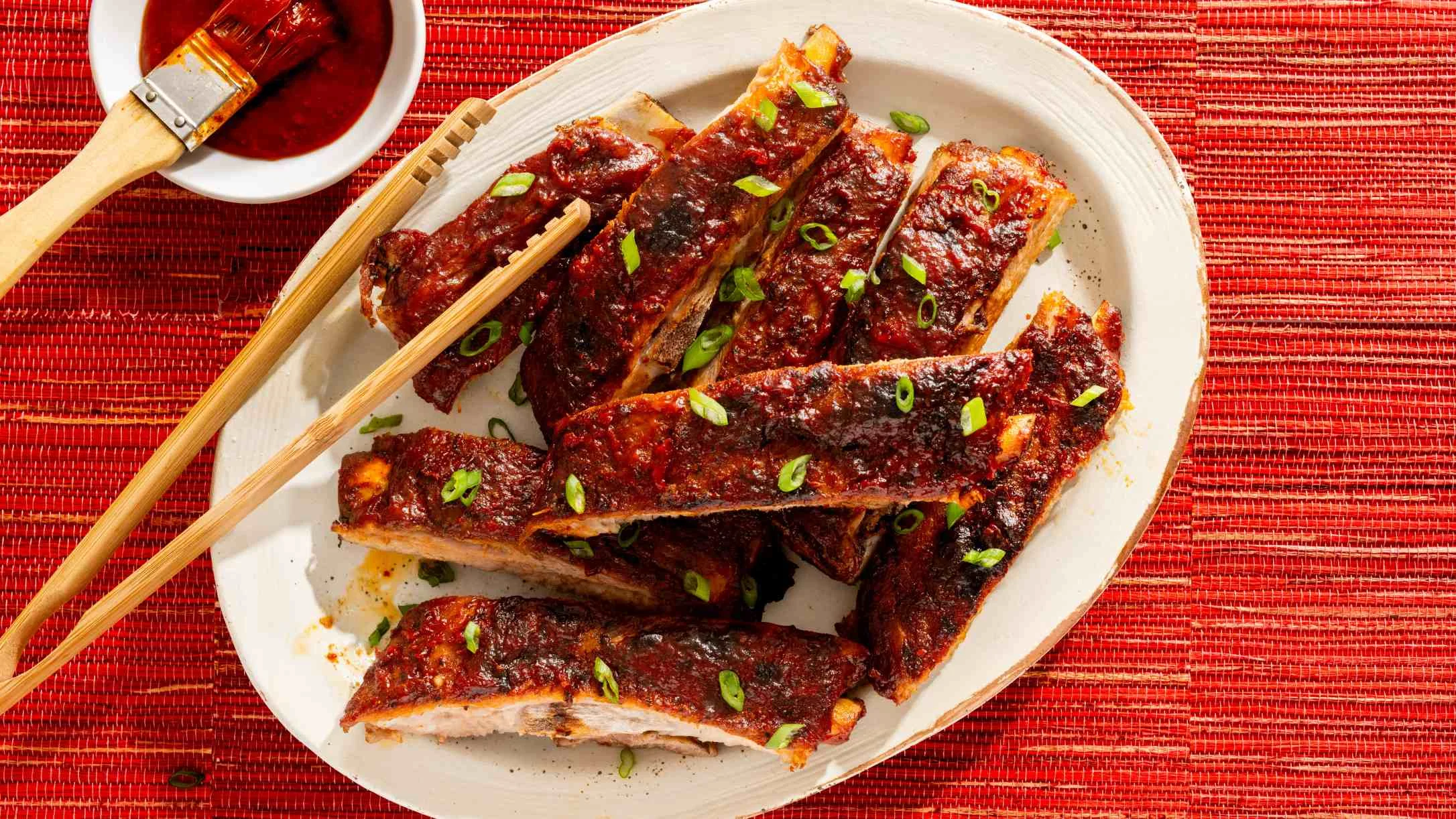 Glazed barbecue ribs garnished with green onions on white plate with sauce on red placemat.