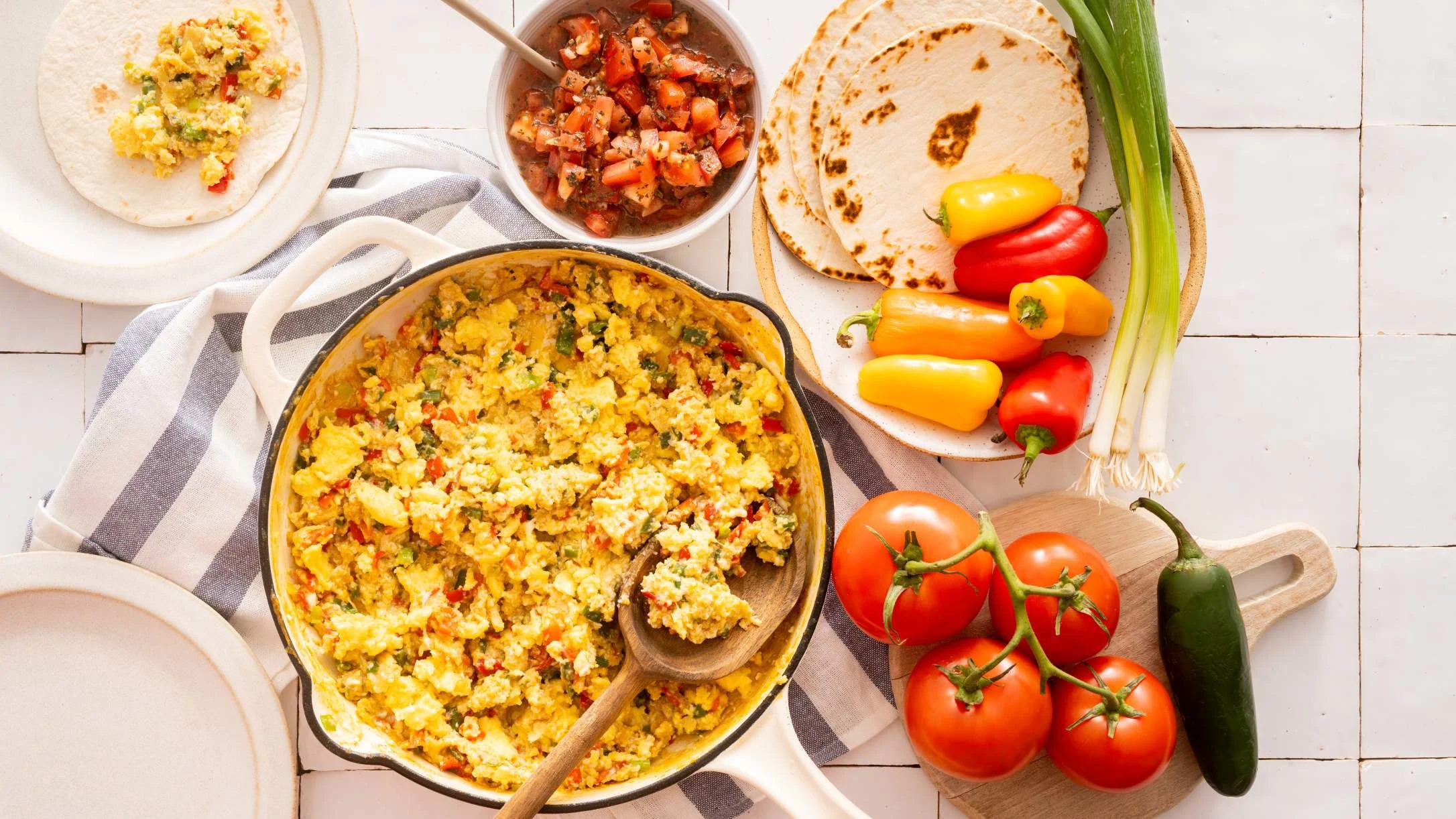 Scrambled eggs with peppers in a yellow skillet, surrounded by tortillas, salsa, tomatoes, mini peppers, and green onions.