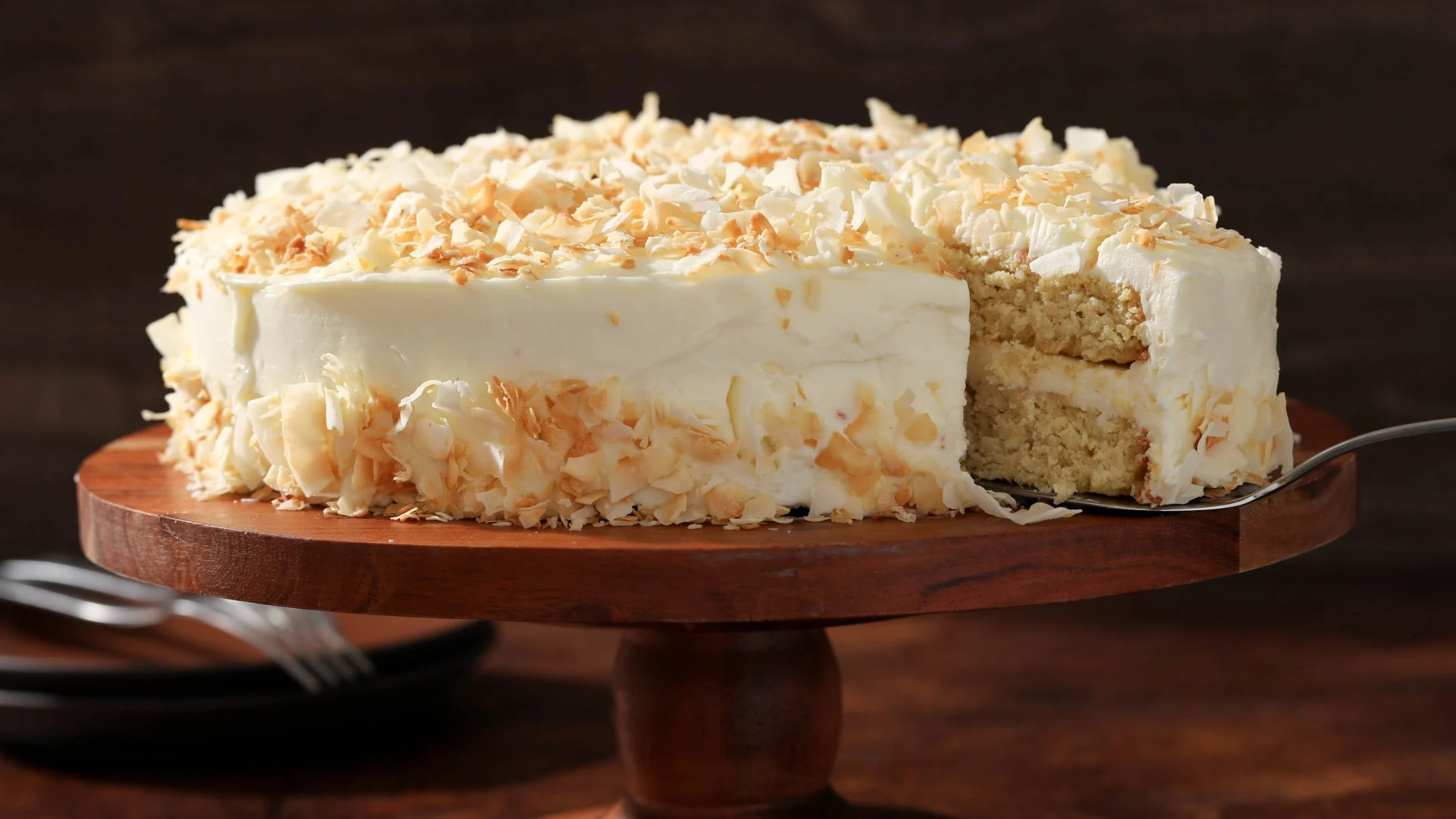 A coconut layer cake with white frosting and toasted coconut flakes on a wooden cake stand, with one slice removed.