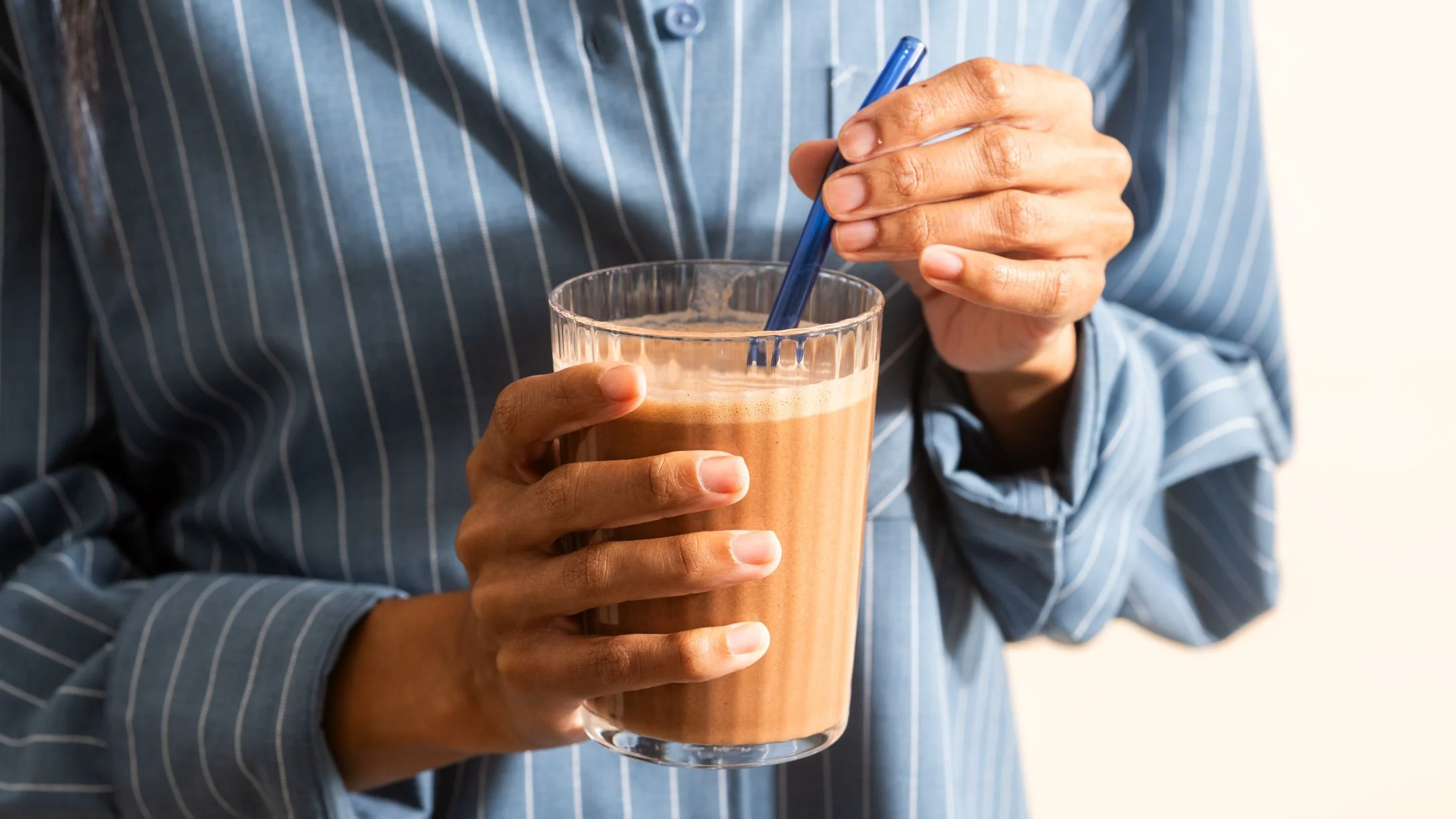 Person in blue striped shirt stirring a glass of chocolate smoothie with a blue straw.