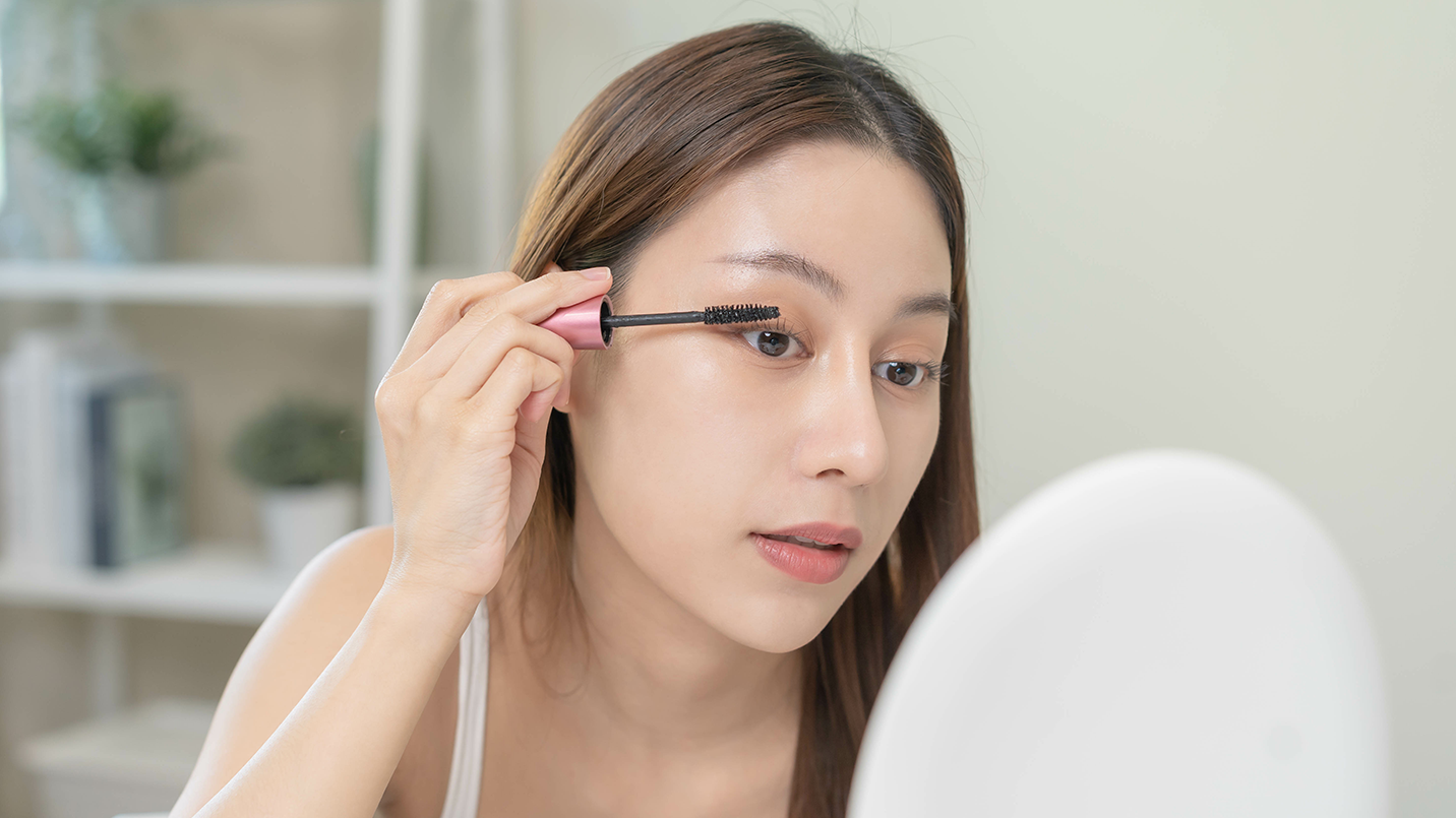 woman applying growth serum to her long eyelashes in front of mirror