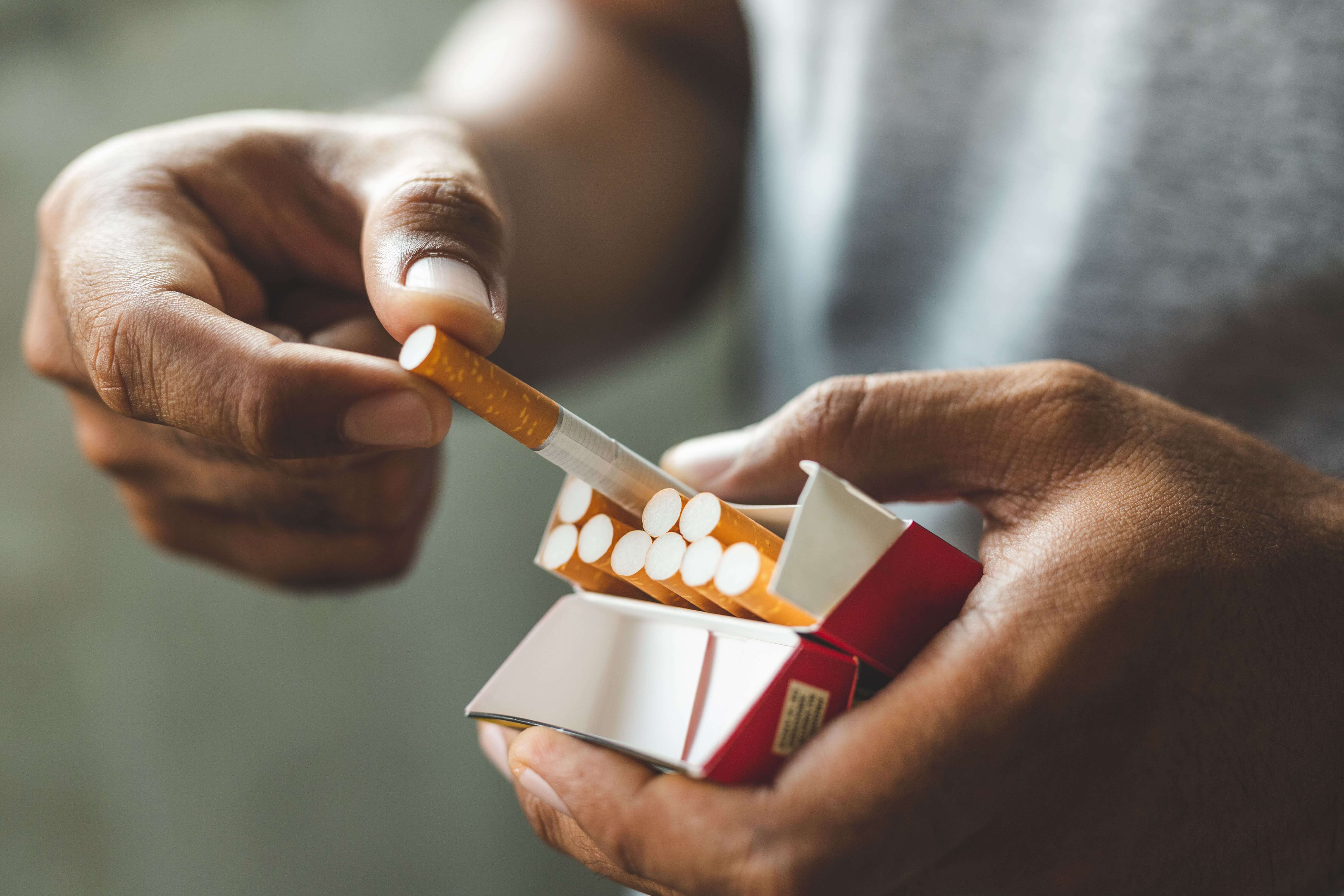 Man with darker skin pulling a cigarette out of a pack of cigarettes.