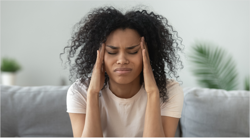 African American woman closing her eyes in pain while holding her head sitting on a sofa