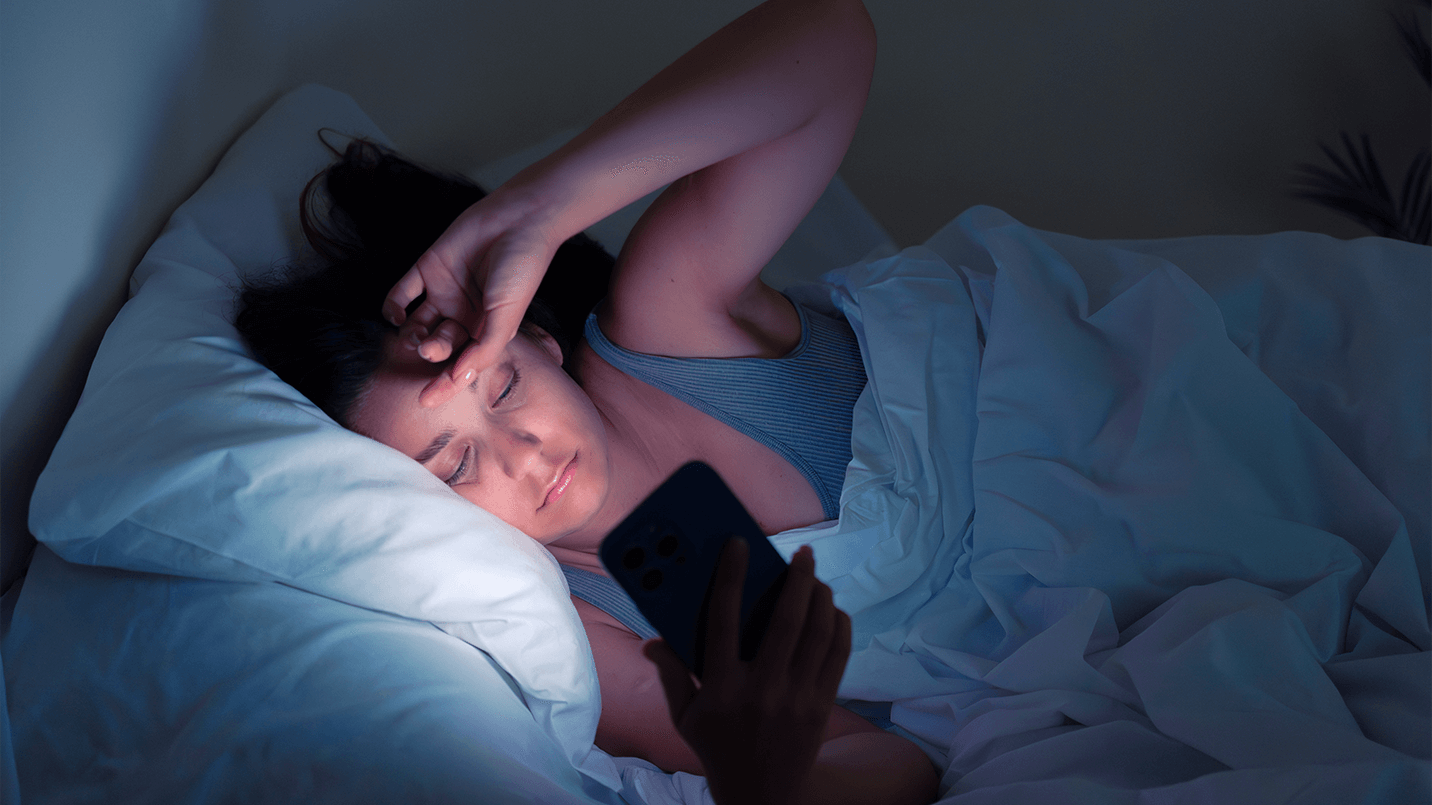 woman in bed looking at her phone in the dark, phone light glowing on her tired face