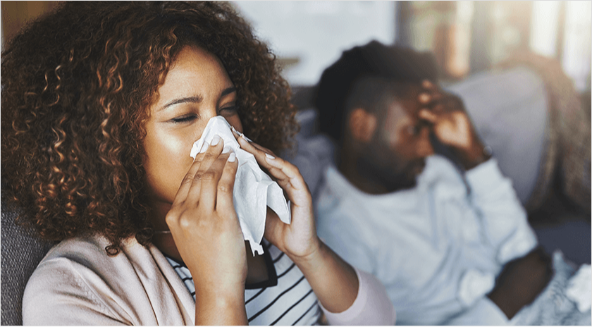 An African American woman and man sitting on a sofa next to each other, while the woman is blowing into tissue.