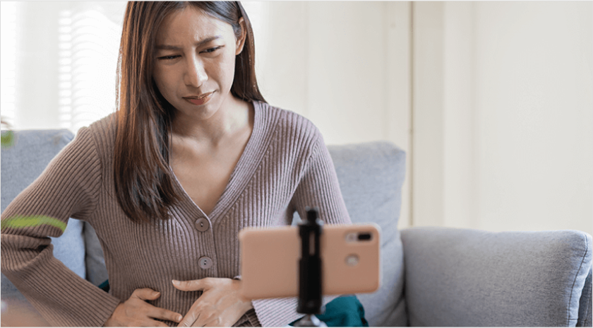 A young woman on a virtual using her phone, who is holding her lower abdomen with a look of pain her on her face 