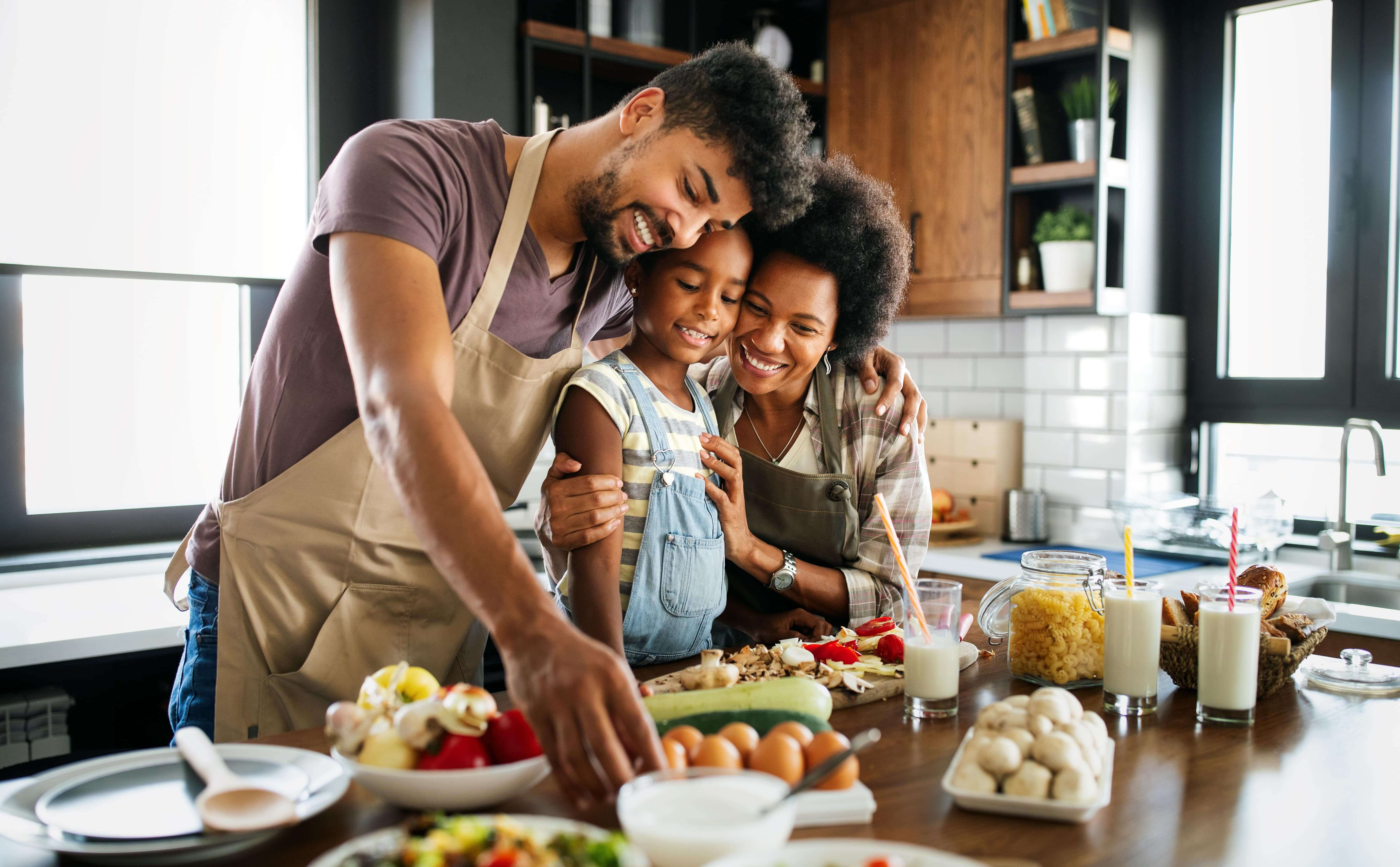 Two adults and their child are making food with healthy ingredients. There are plenty of healthy and whole ingredients on the table and the family is hugging together. 